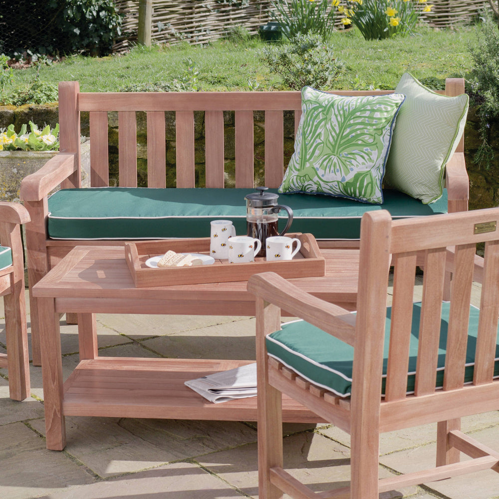 
                  
                    Teak outdoor chairs and teak bench with green seat pads and  thick slatted backrest, smooth curved armrests, behind teak coffee table with shelf, holding teak tray with coffee pot and cups. 
                  
                