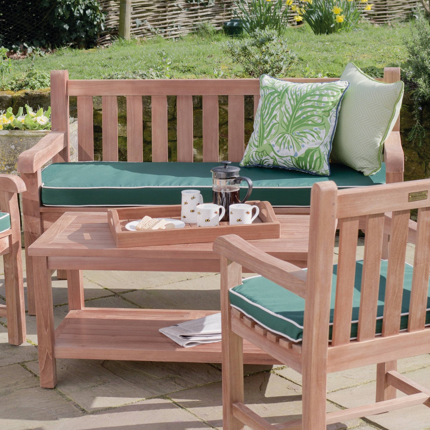 Teak outdoor chairs and teak bench with green seat pads and  thick slatted backrest, smooth curved armrests, behind teak coffee table with shelf, holding teak tray with coffee pot and cups. 