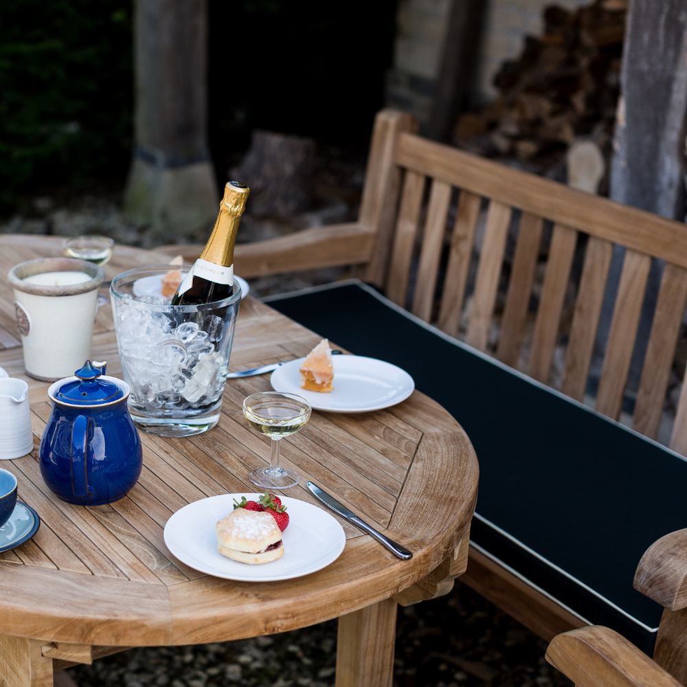 
                  
                    Teak bench with armrests and backrest, with dark green seat cushion, in front of oval teak dining table laid with teapot, wine cooler, plates and candle. 
                  
                