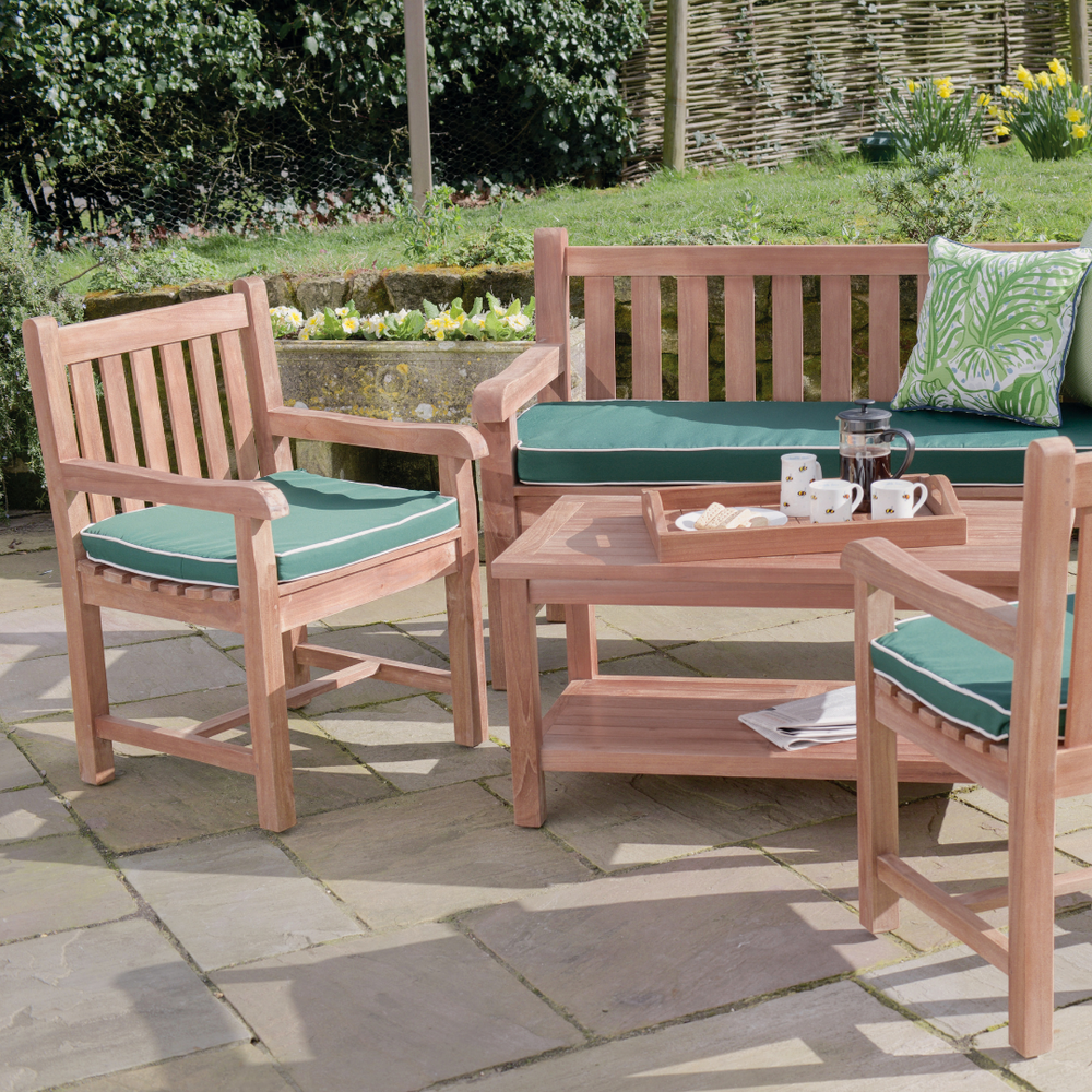 
                  
                    Teak outdoor bench and teak chairs with armrests, all with green seat cushions, around teak coffee table with tray of coffee and cups, in garden setting. 
                  
                