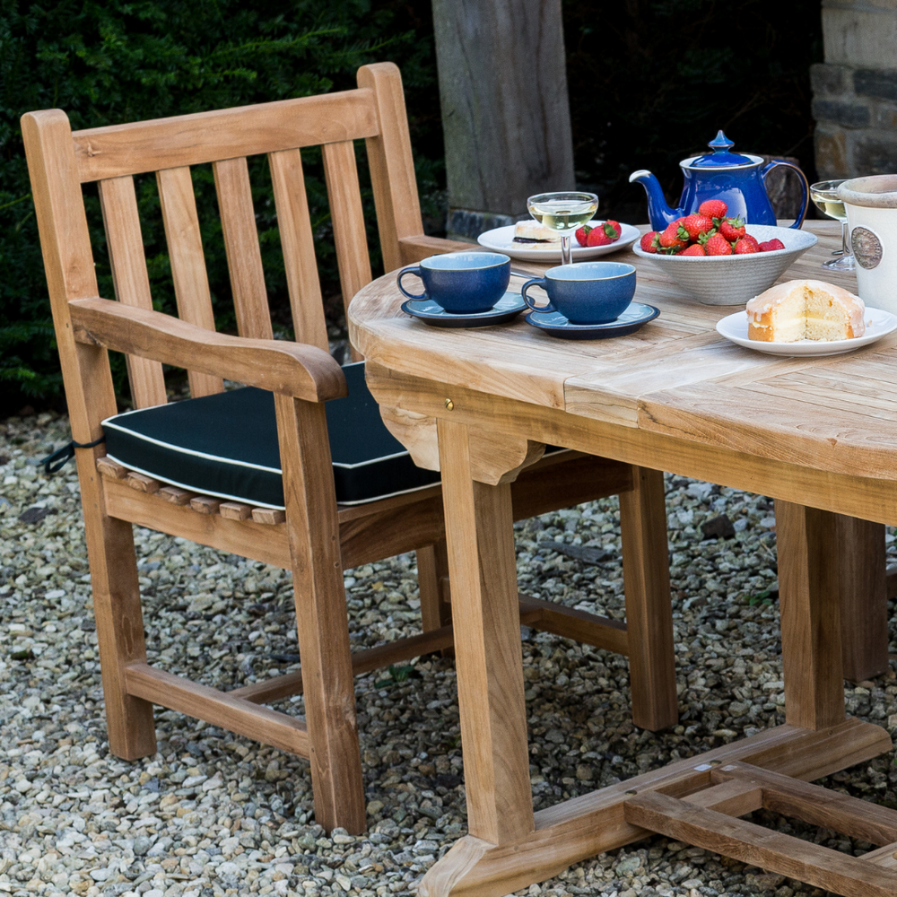 
                  
                    Single teak dining chair with armrests, next to oval teak dining table with blue tea set, bowl of strawberries and cake on white plate, on gravel patio. 
                  
                