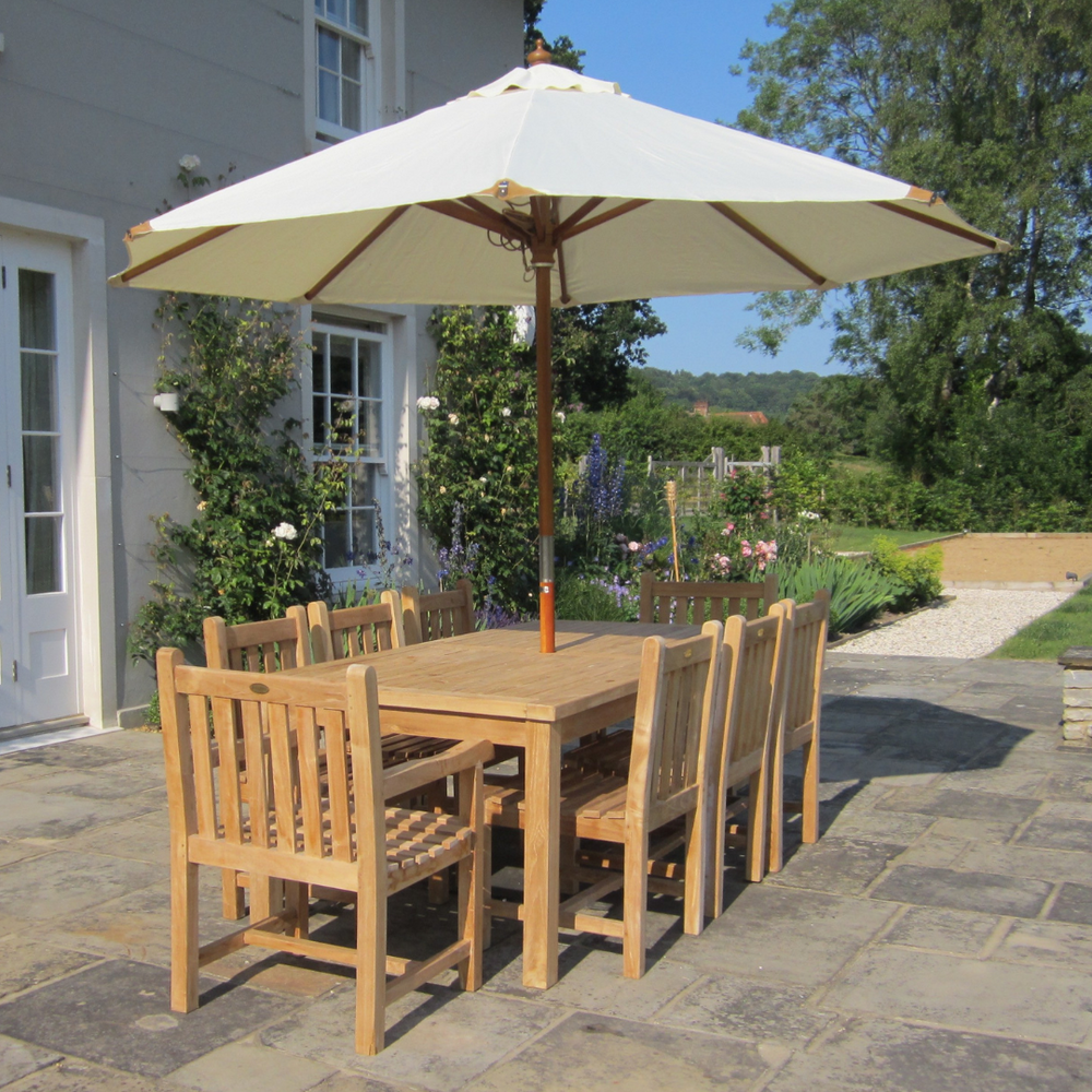 
                  
                    Teak rectangular dining table with large white parasol and eight teak dining chairs, on stone patio with garden, flowerbeds and house in background
                  
                