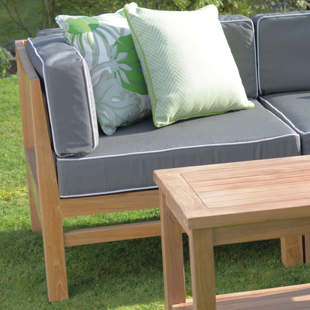 
                  
                    Teak outdoor sofa with grey and white seat and back cushions and green patterned scatter cushions. In front is a teak coffee table, on a grassy surface 
                  
                