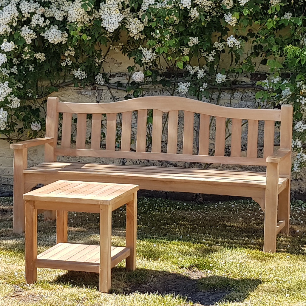 Square teak coffee table in front of teak bench with curved backrest, in a garden setting. 