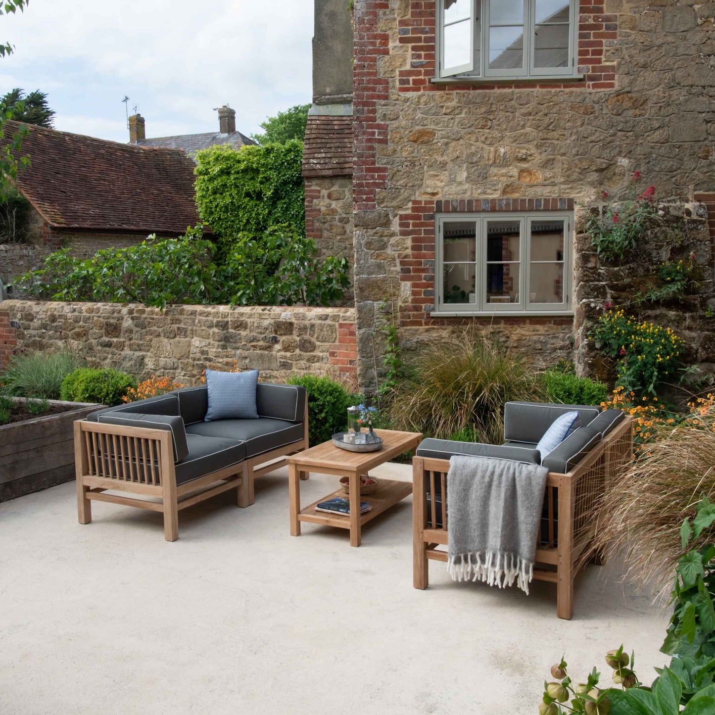 Teak rectangular coffee table with shelf, with teak benches either side, with grey-green cushions, on stone patio. Behind is imposing brick and stone house.