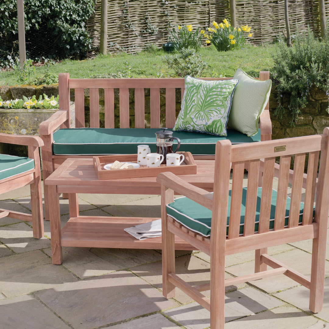 Teak outdoor bench and teak chairs with armrests, all with green seat cushions, around teak rectangular coffee table with tray of coffee and cups, in garden setting. 