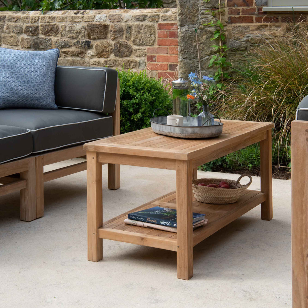 Teak  Rectangular Coffee Table with tray with flowers, jug and lantern, next to teak outdoor sofa with dark grey and white cushions, on a stone patio. 
