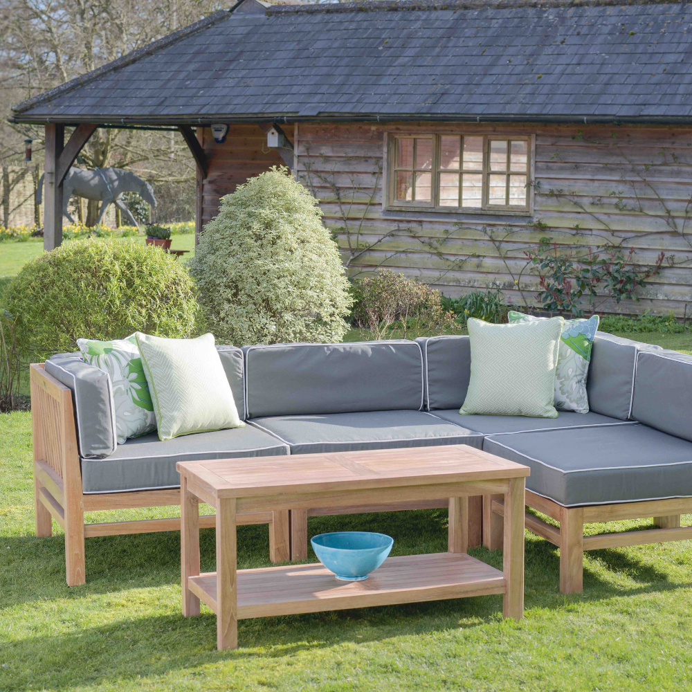 Teak Rectangular Coffee Table in front of teak corner sofa with green - grey seat and backrest cushions and green scatter cushions, in a garden with a wooden building in the background 