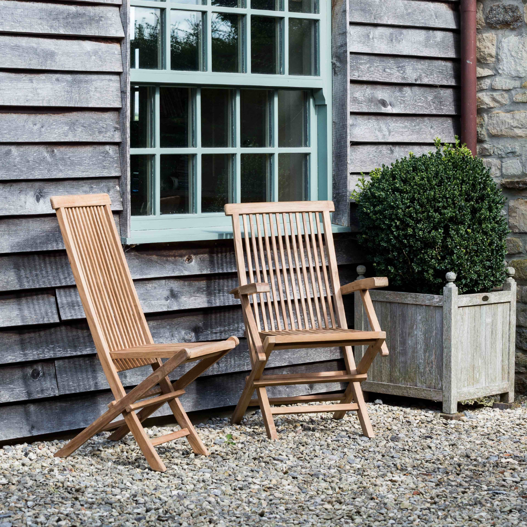 Two Folding Dining Teak Chairs, one with armrests, one without, in front of wooden building, on gravel patio. 