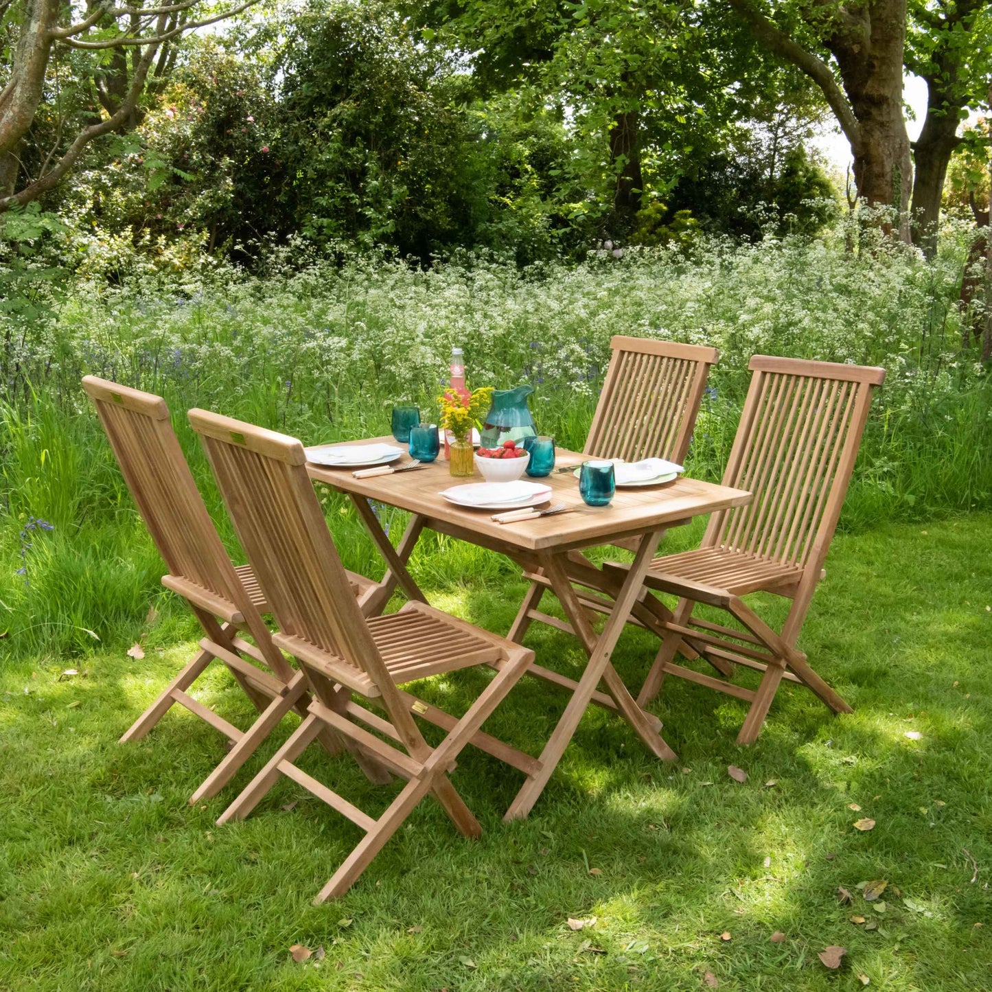 Four teak folding dining chairs around teak rectangular dining table with place settings and glasses, in dappled sunshine, in garden setting