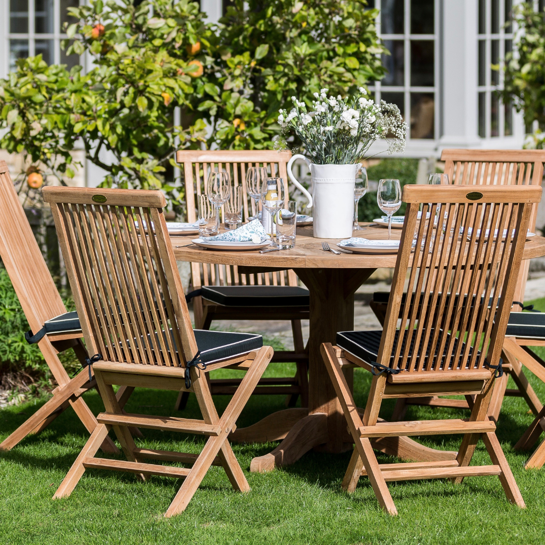 Five teak folding dining chairs around round teak dining table, laid with glasses and plates, with white jug of flowers in centre, in garden setting in front of house with bay windows