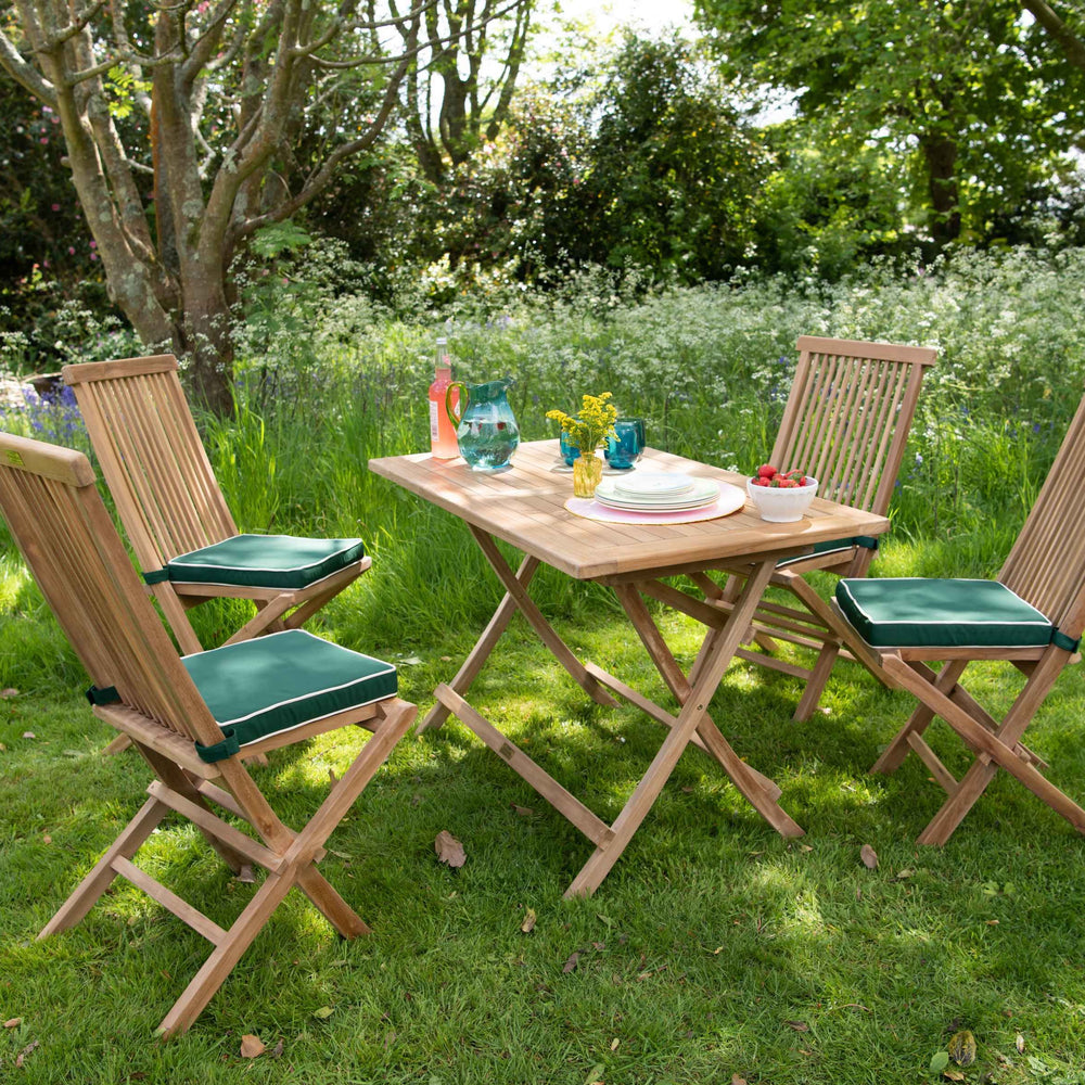 
                  
                    Teak folding dining chairs with green cushions, set at angles around teak folding rectangular dining table with green jug and glasses, in sunny garden
                  
                