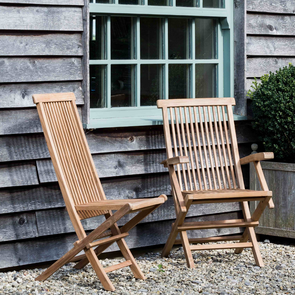 
                  
                    Two teak folding dining chairs, side by side. One with armrests, one without, on gravel patio with wooden building behind. 
                  
                