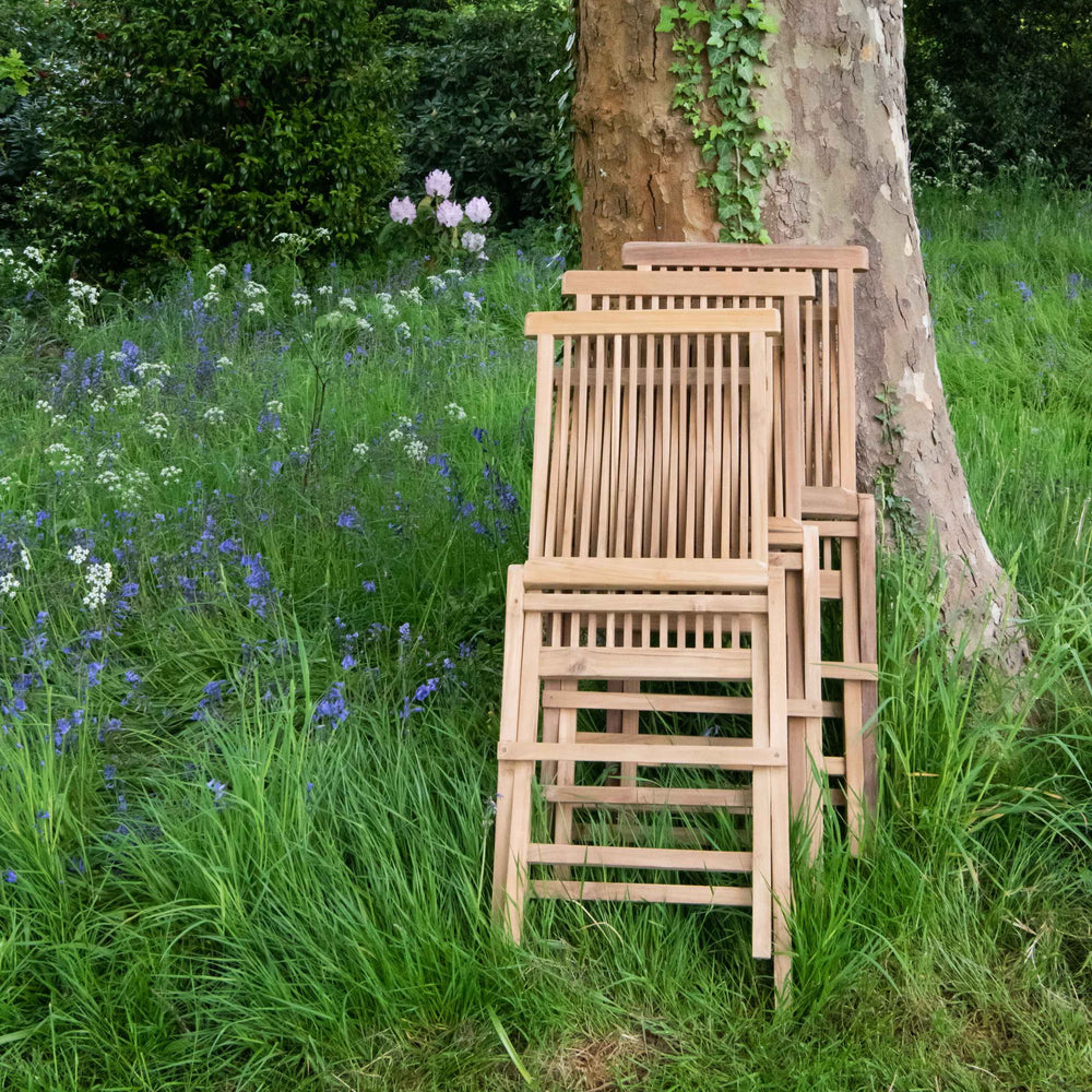 
                  
                    Three teak folding dining chairs, in folded position, stacked against tree, in garden setting with wild floweres.
                  
                