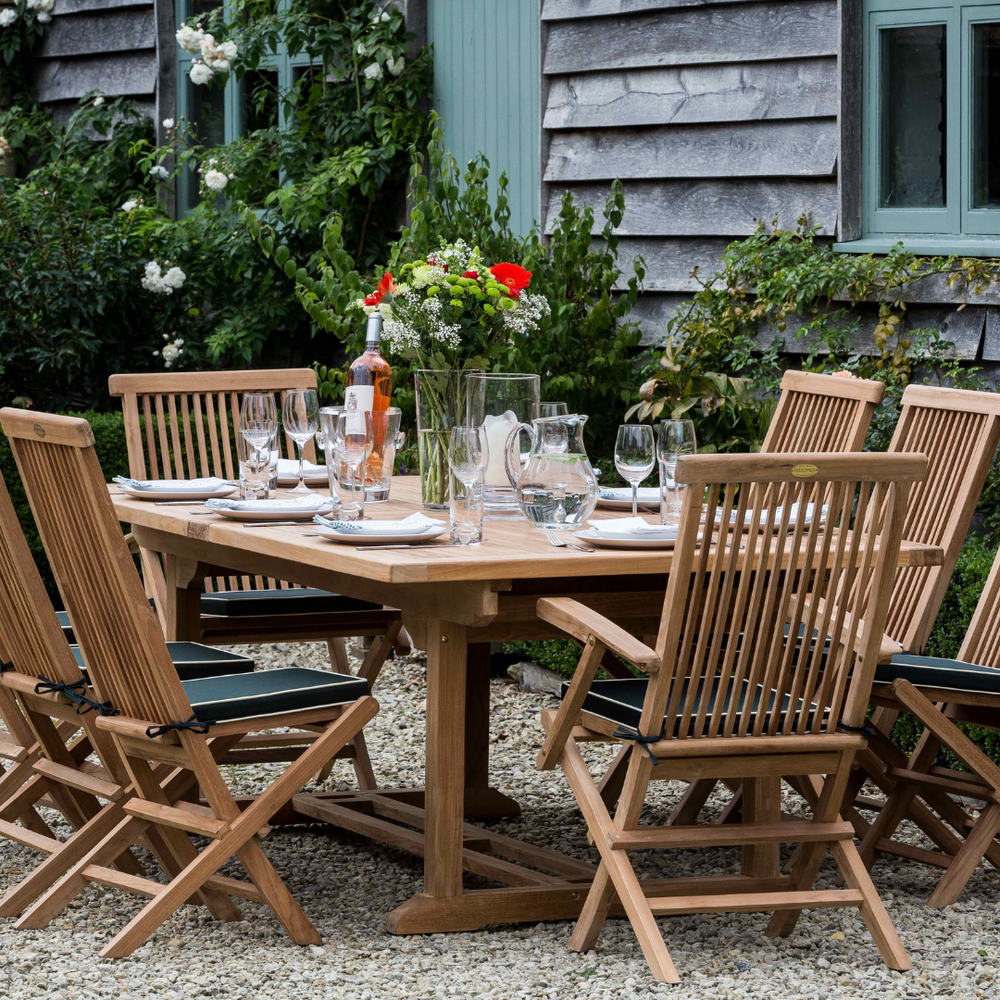 
                  
                    An extendable rectangular teak dining table set with teak dining chairs with green cushions, laid out for a meal with plates, glasses, and a floral centerpiece, on an outdoor patio.
                  
                