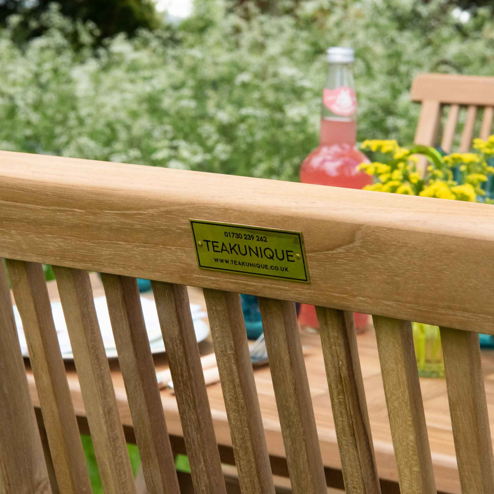 
                  
                    Close up of back view of teak folding dining chair with small brass plate screwed displaying word Teakunique. In background is a teak table with flowers and bottles
                  
                