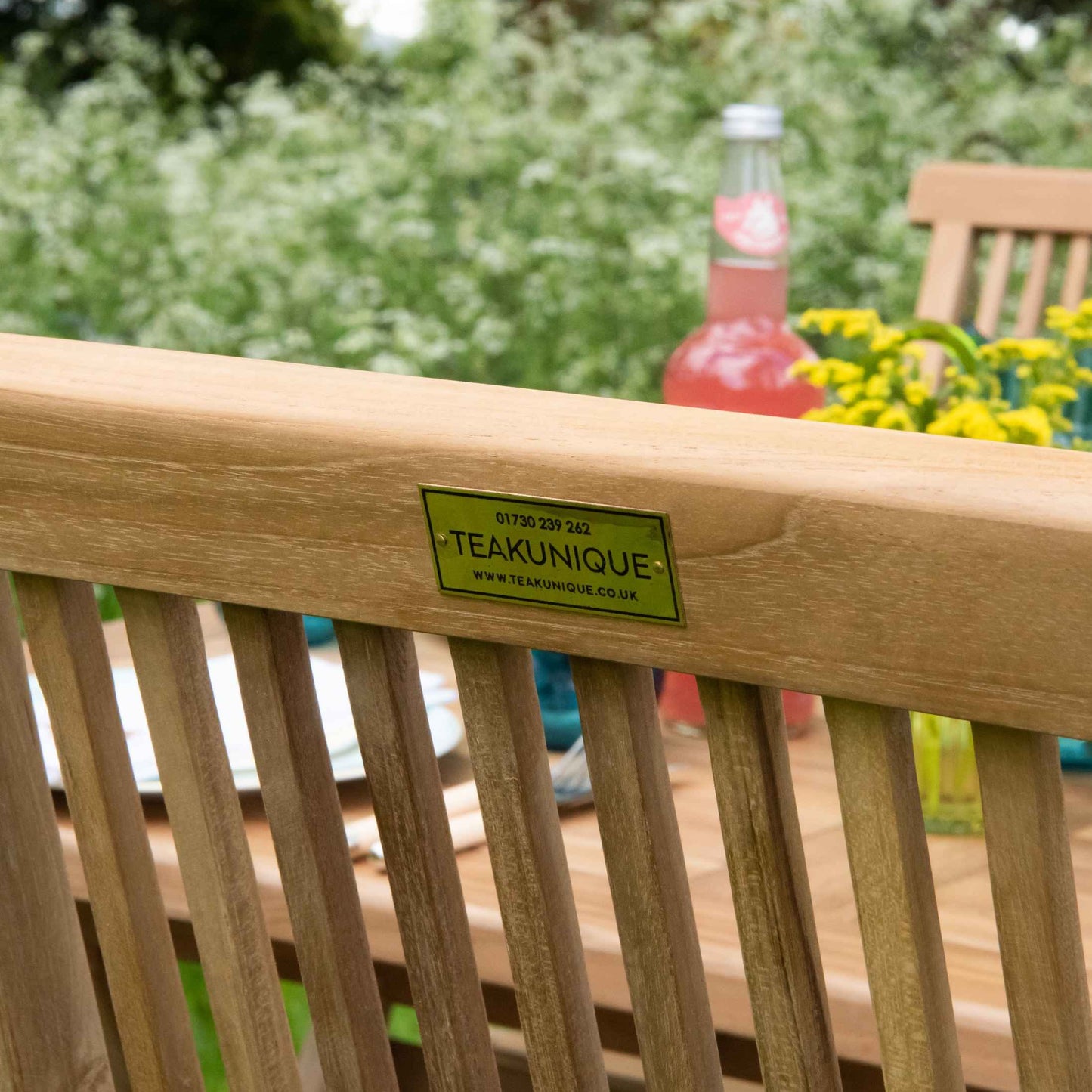 Close up of back view of teak folding dining chair with small brass plate screwed displaying word Teakunique. In background is a teak table with flowers and bottles