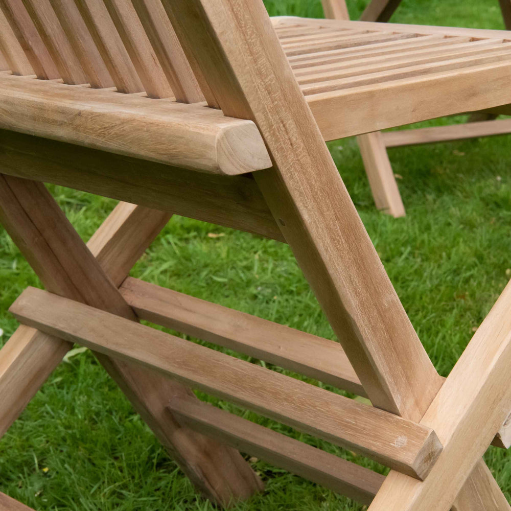 
                  
                    Close up of teak folding dining chair, with focus on hinge between slatted seat and slatted backrest, on grassy lawn. 
                  
                