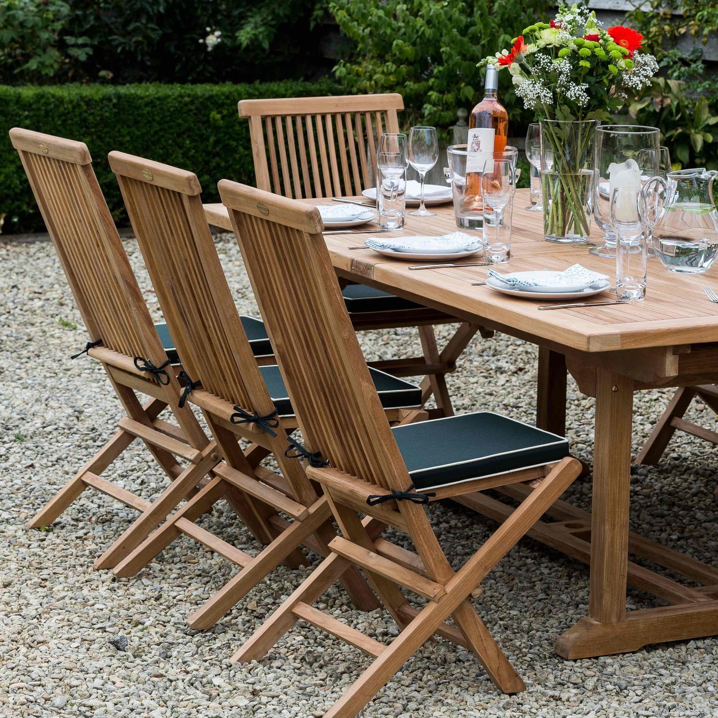 Teak rectangular dining table on a gravel surface. The table is set with place settings, glasses and flowers, and four teak folding dining chairs with green seat cushions. 