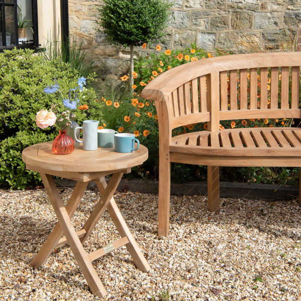 
                  
                    Teak crescent-shaped bench with slatted seat and backrest, next to small round folding teak table set with cups and flowers, on gravel patio with flowerbeds behind. 
                  
                