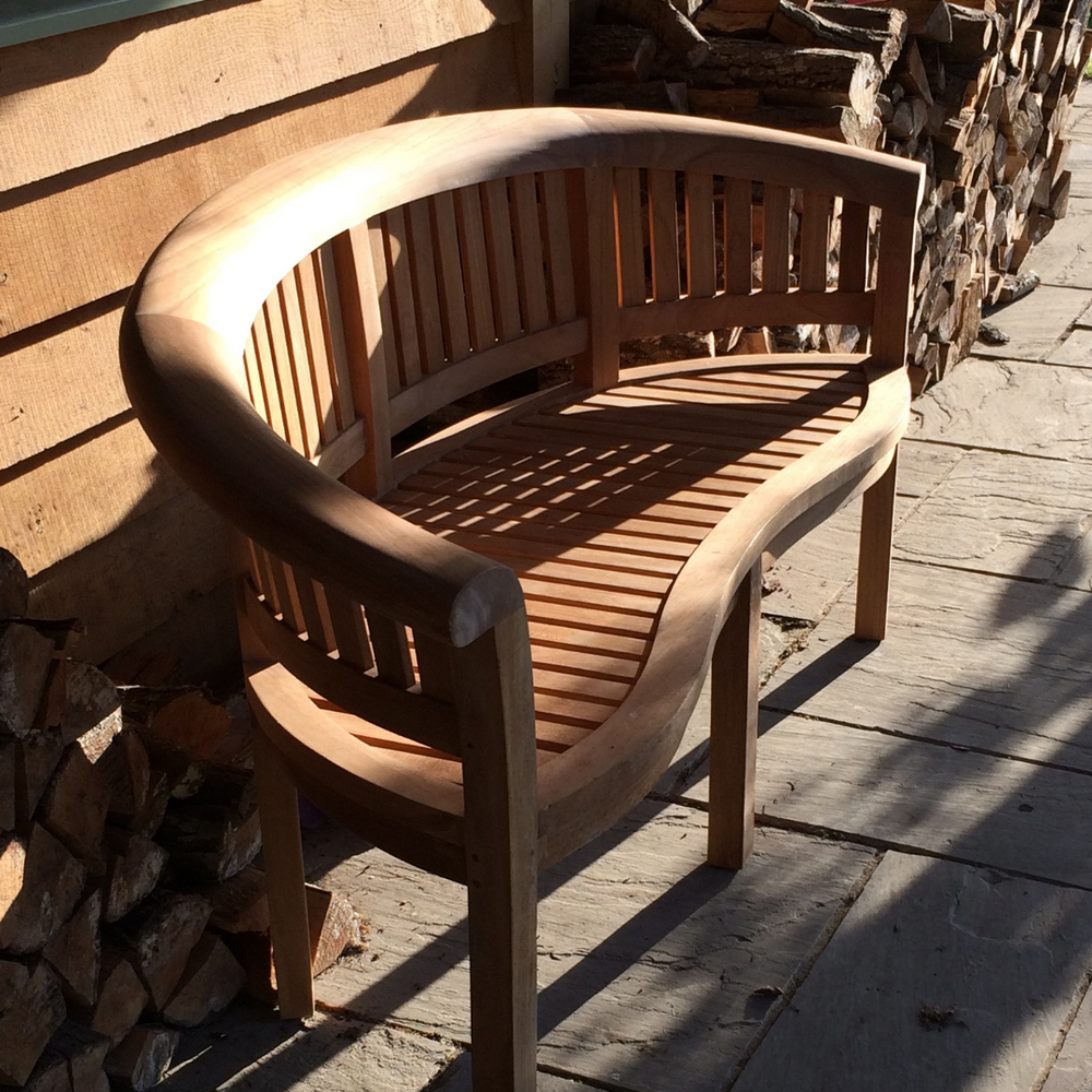 
                  
                    Teak crescent-shaped bench with curved seat, on wooden floor next to piles of logs, in front of wooden building. 
                  
                