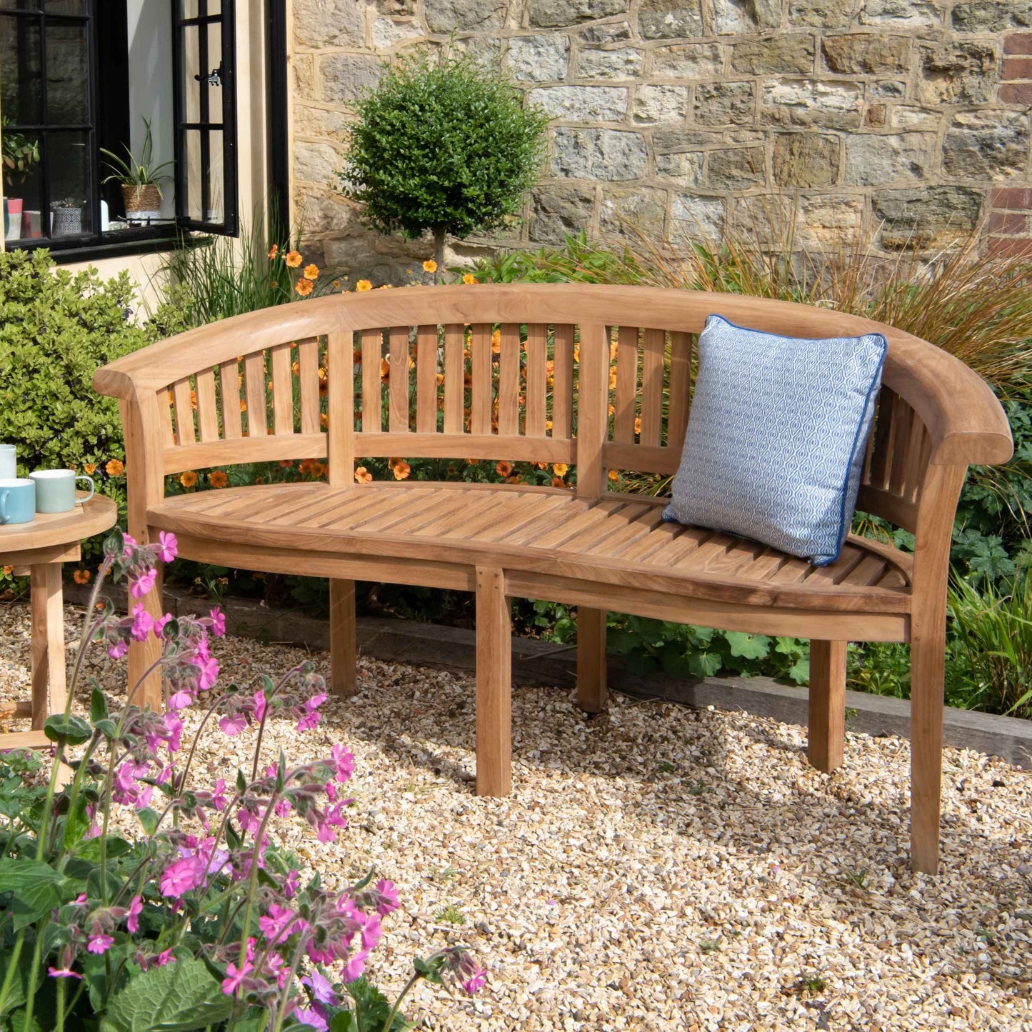 Teak crescent-shaped bench with blue patterned scatter cushion, next to small round teak table with cups, on gravel patio with stone wall and open window in background