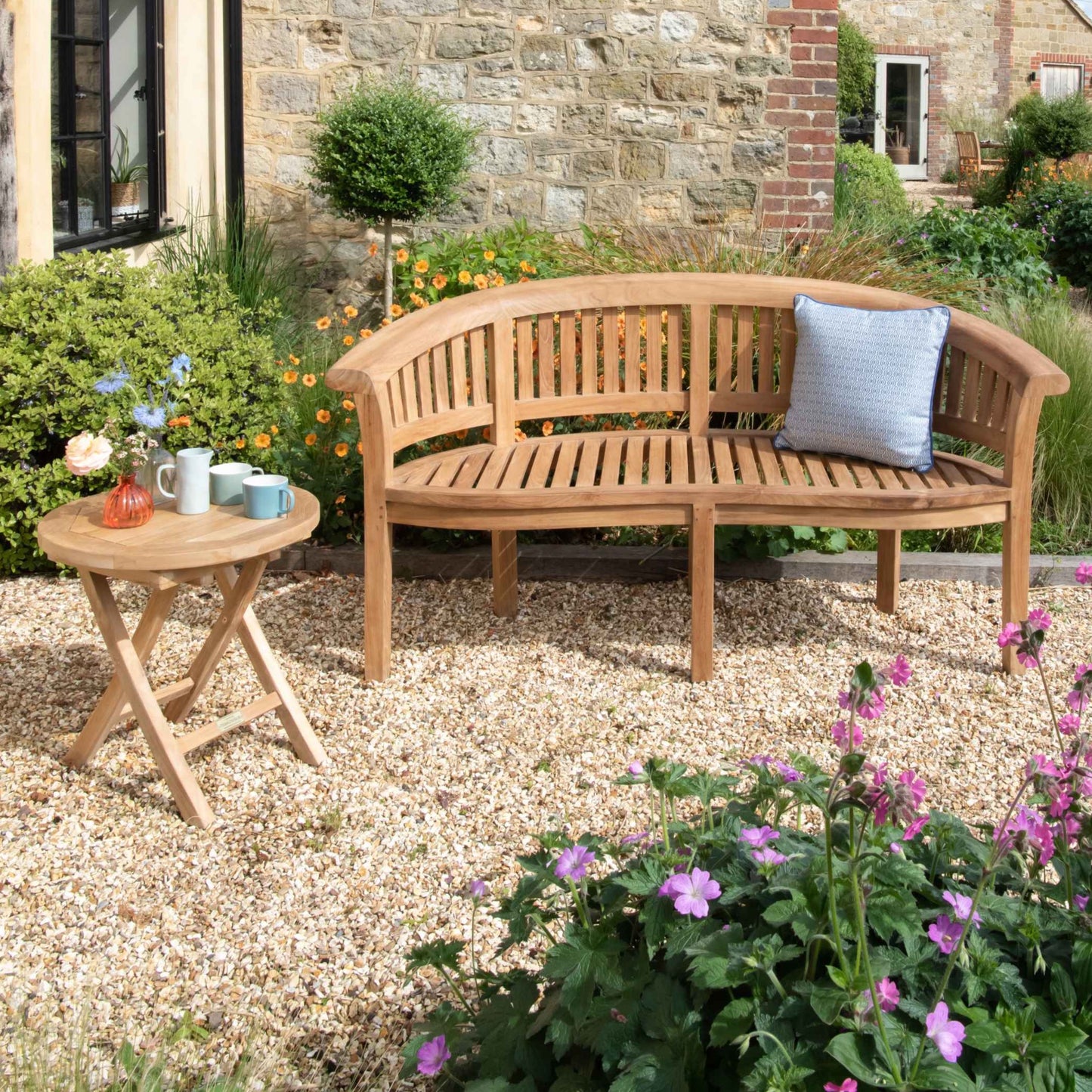 Teak crescent shaped bench with blue patterned cushion, next to small round folding teak table with cups and flowers, on a gravel patio with brick and stone house in the background