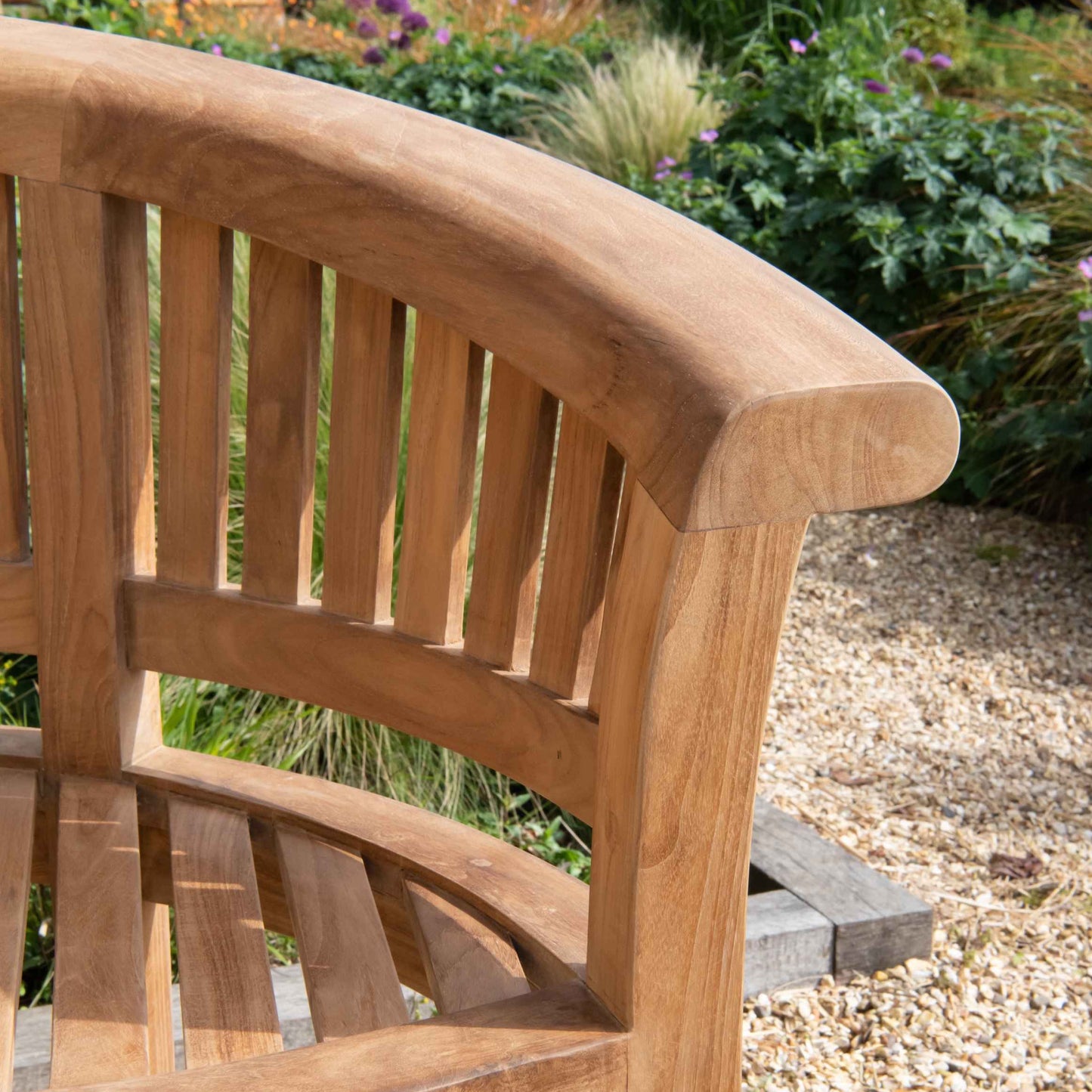 One end of a teak crescent-shaped bench on a gravel surface with shrubs behind. 