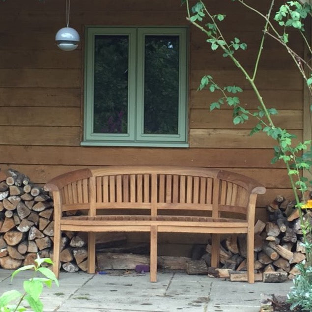 Teak crescent shaped bench in front of wooden wall with green painted window. To both sides of the bench are piles of logs.  