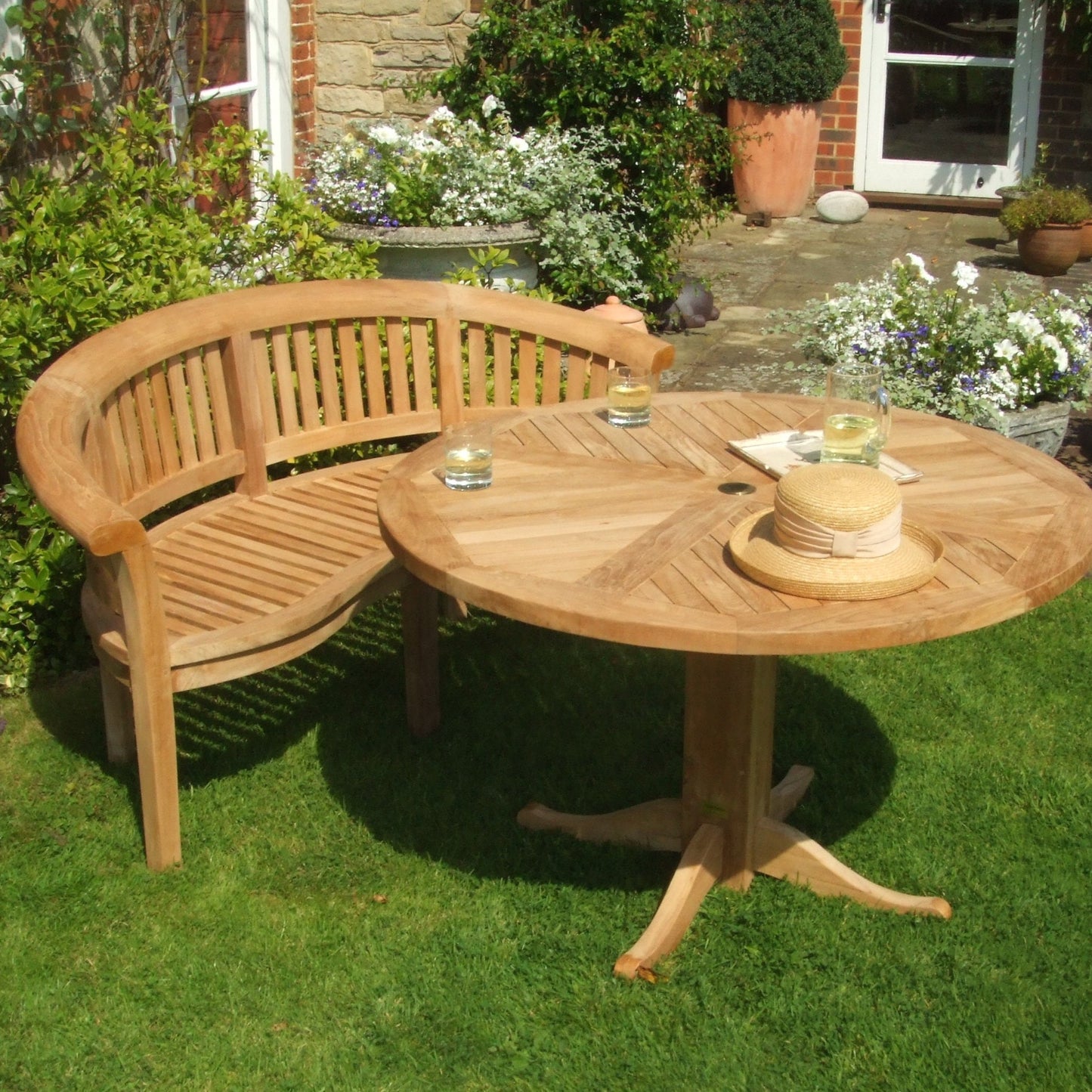 Teak crescent shaped bench and teak round pedestal table, holding glasses and a straw hat, on grassy surface and in front of a brick and stone building