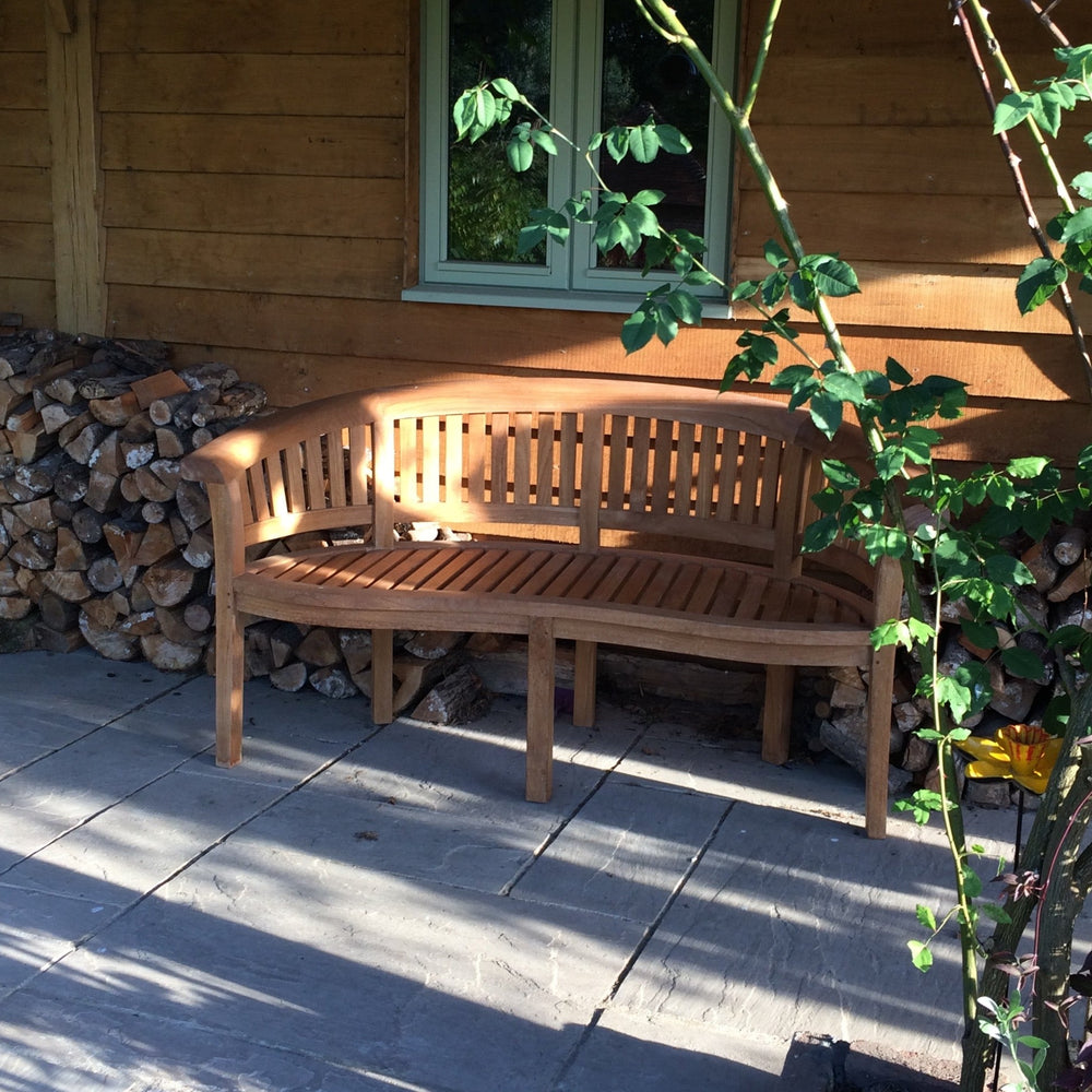 
                  
                    Teak crescent shaped bench on stone patio, with log piles either side, in front of wooden wall with green window. 
                  
                