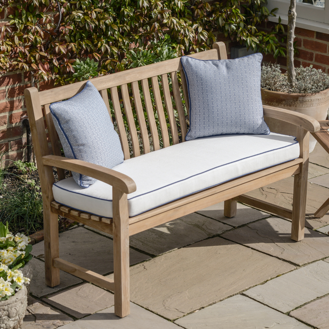 Teak bench with slatted backrest and curving armrests, with cream coloured seat cushion and blue scatter cushions, on patio with ceramic planters.  