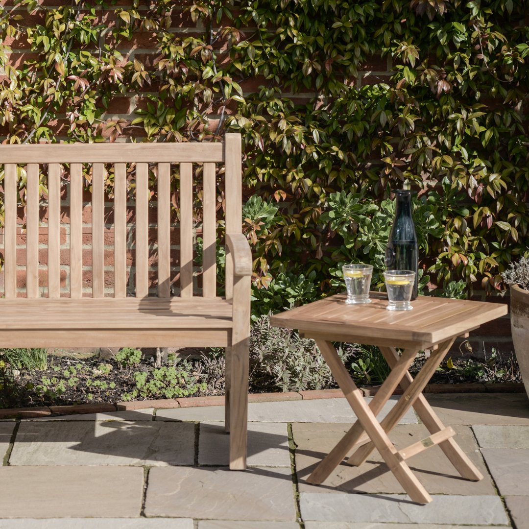 Teak outdoor bench next to small square teak folding side table, with water glasses and bottle, on stone patio with hedge in background 