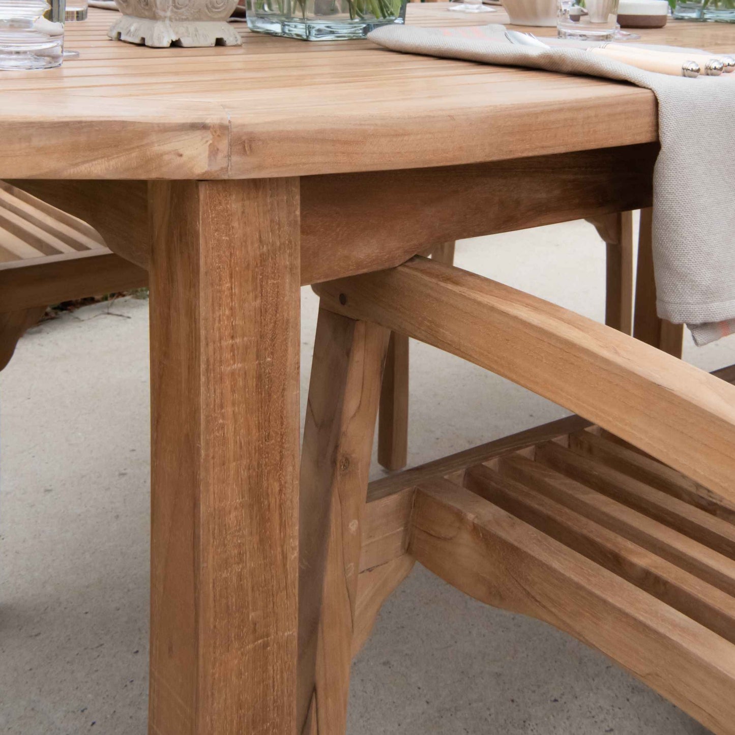 Close up of corner of teak outdoor dining table with teak armchair tucked underneath. On the table is a napkin and cutlery. 