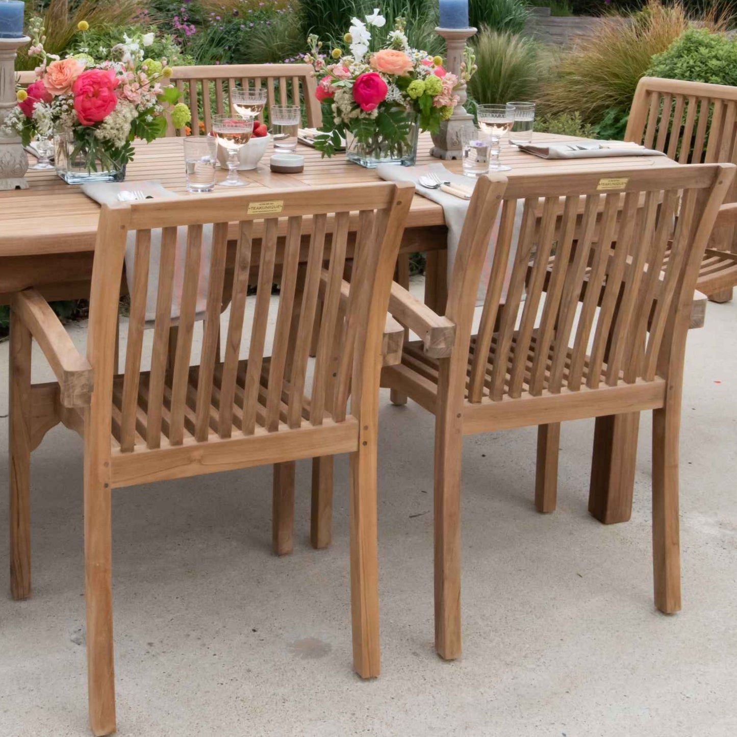 Teak outdoor dining table and armchairs on a stone patio. The table has flower arrangements, glasses and place settings.  