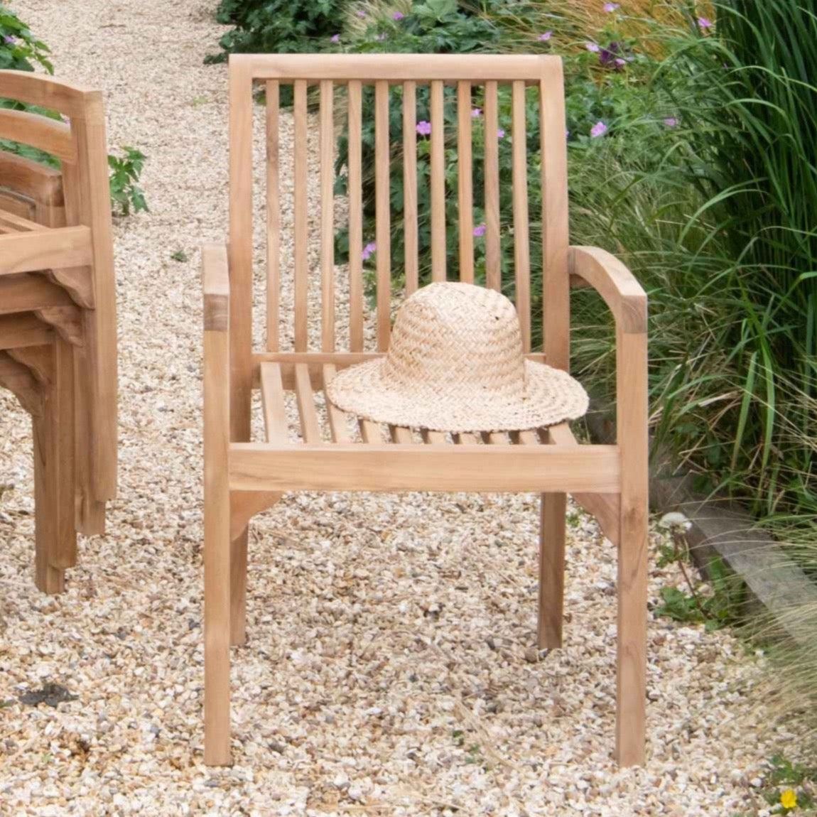 A teak slatted stacking carver chair with a straw hat placed on the seat, set on a gravel surface with plants in the background.