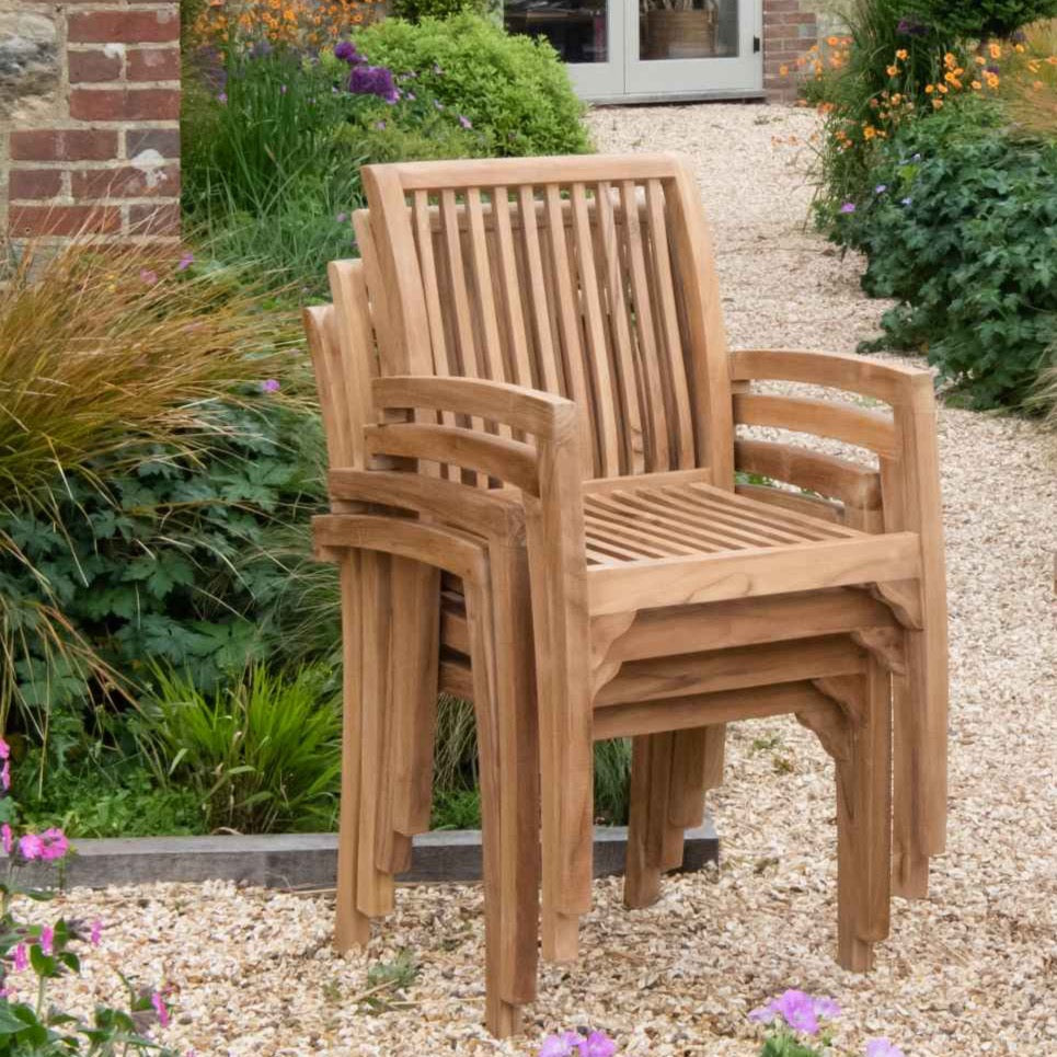 
                  
                    Stack of four teak slatted carver chairs on a gravel patio with greenery and flowerbeds behind 
                  
                