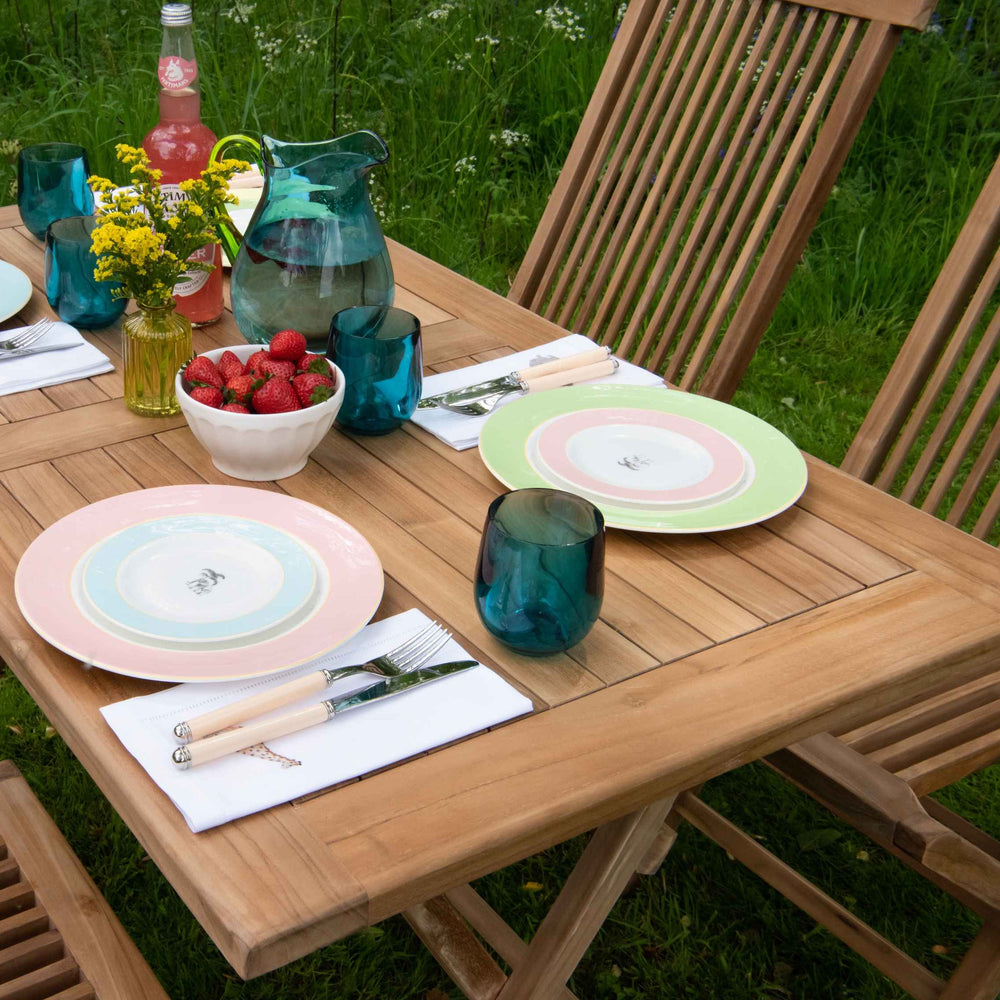 
                  
                    Rectangular folding teak dining table in garden, with teak dining chairs with slim slatted seat and backrest. The table holds pastel coloured plate settings, fruit and water jugs and glasses.
                  
                