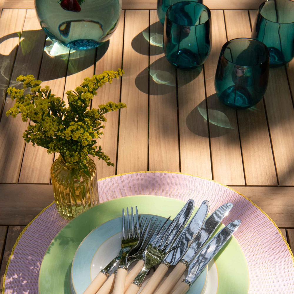 
                  
                    Close up of tabletop of teak folding rectangular table with green glasses and pastel coloured plates and flowers. 
                  
                