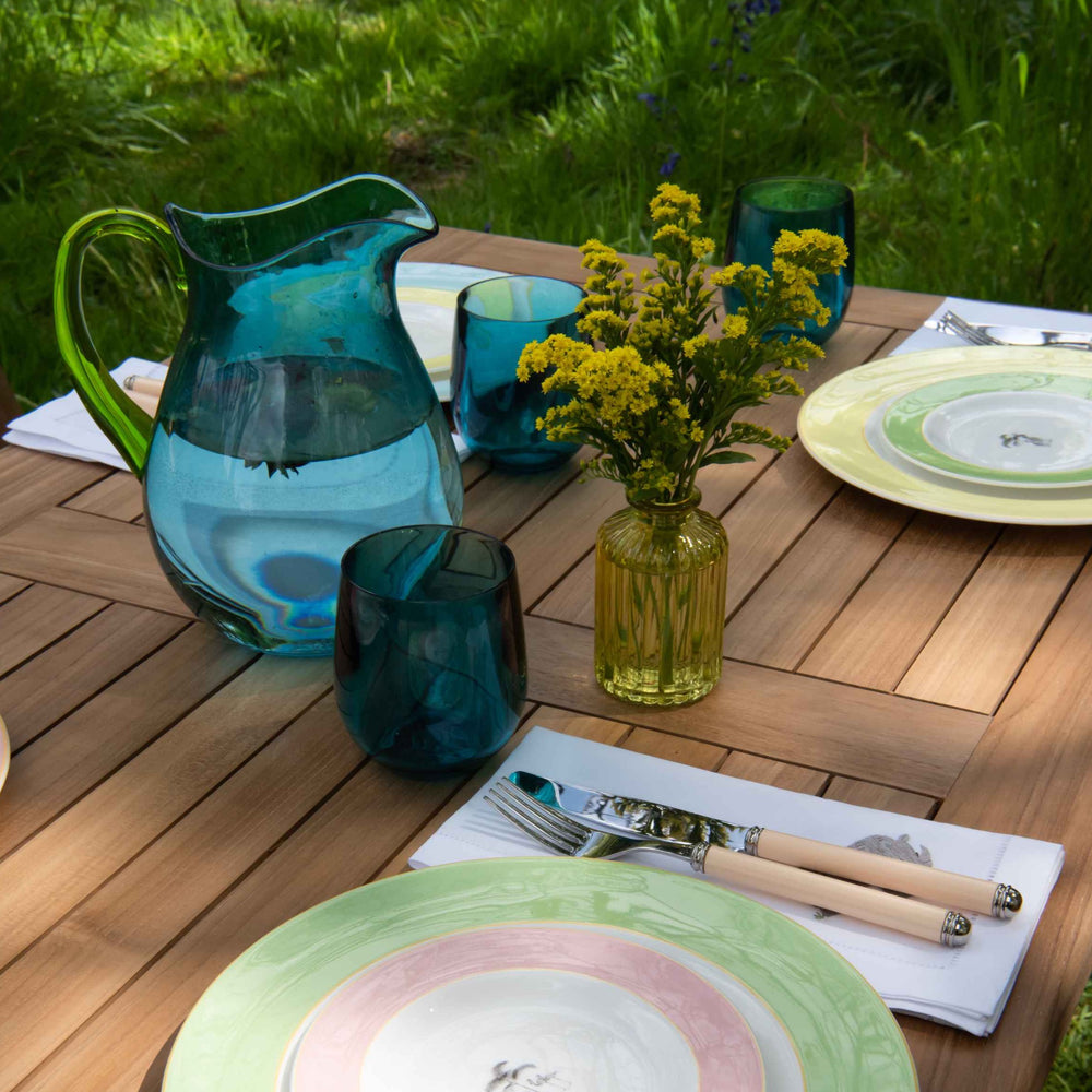 
                  
                    Close up of tabletop of teak folding rectangular table with  pastel coloured plates and cutlery, green glasses and jug and small vase of flowers. 
                  
                