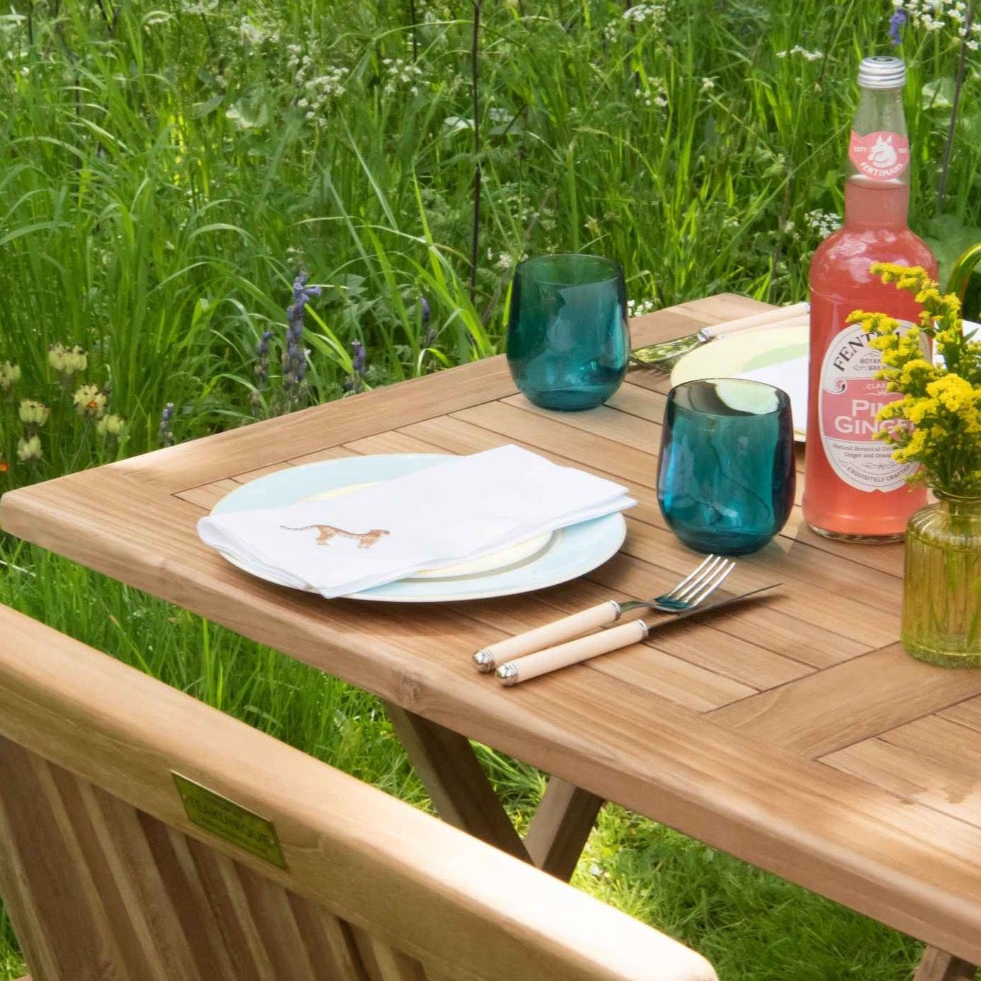 Teak folding rectangular table set with plate and cutlery, glasses and bottles. There is grass and wildflowers in the background