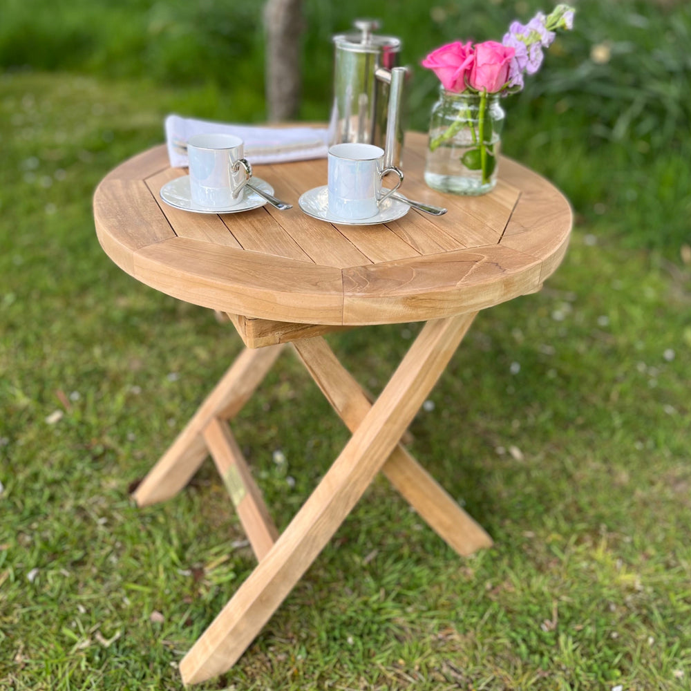 A round folding teak coffee table on a grassy area, with a teacup, saucer, and a vase with flowers on top of it.