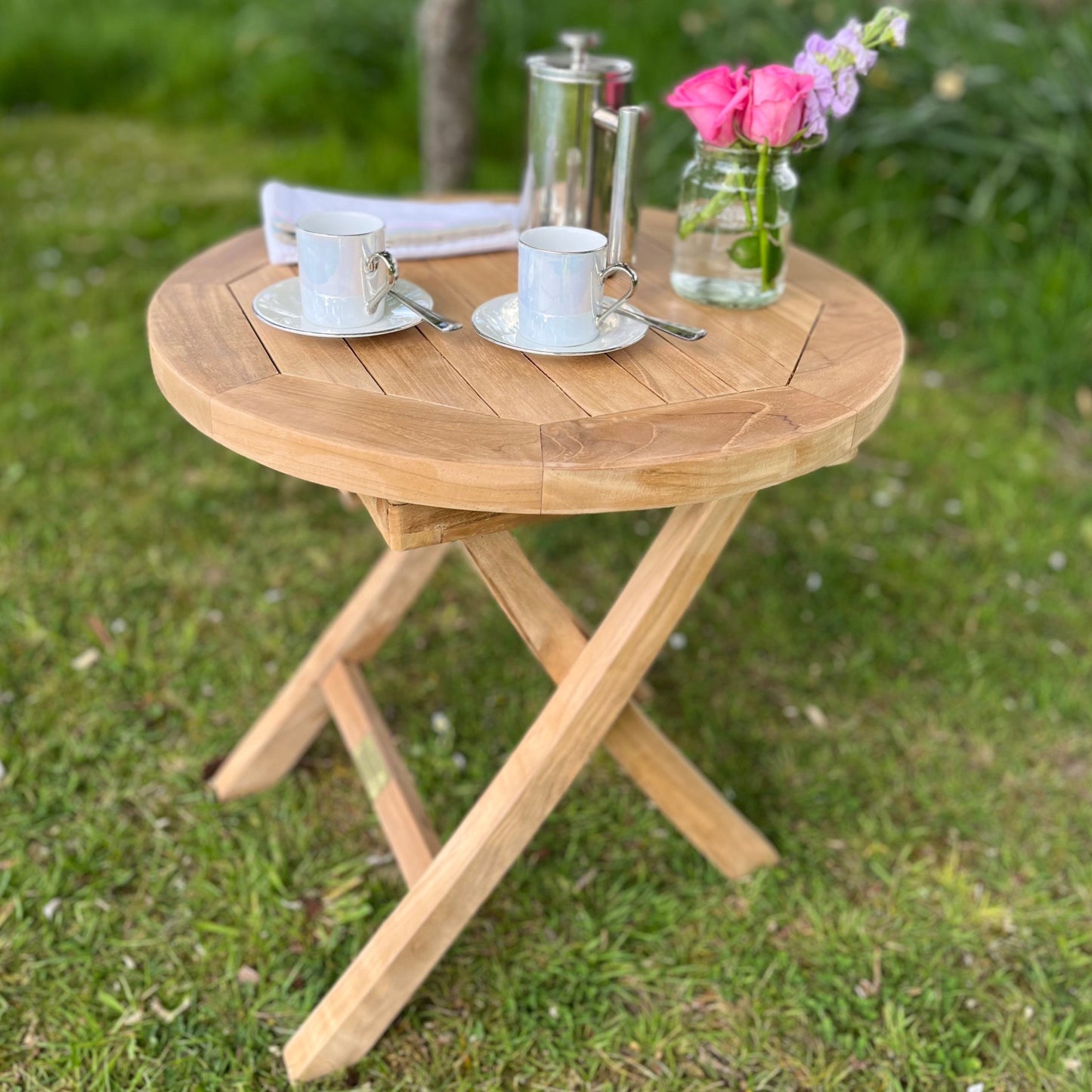 A round folding teak coffee table on a grassy area, with a teacup, saucer, and a vase with flowers on top of it.