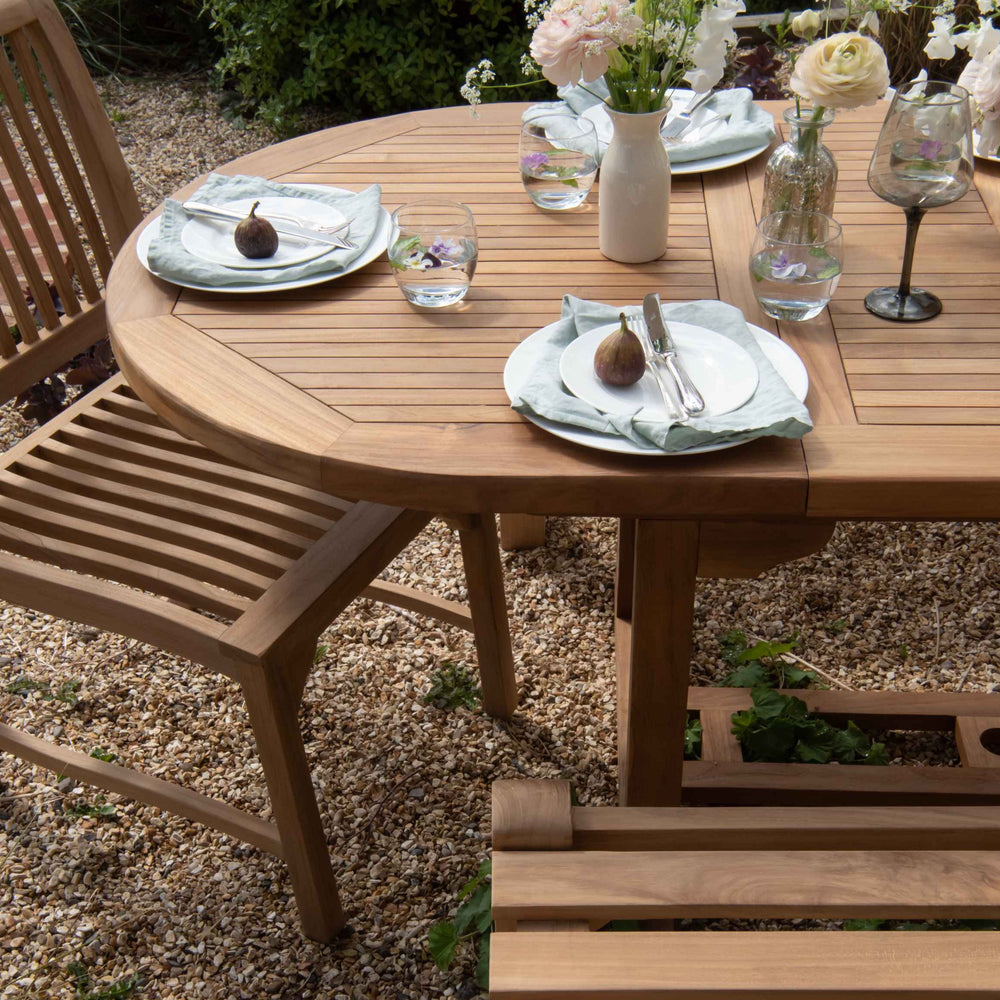 
                  
                    Teak oval dining table with teak bench and dining chair set around, with white plates, wine glasses and vases of flowers, on gravel patio. 
                  
                