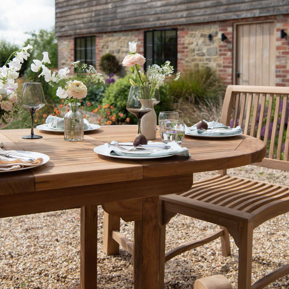 
                  
                    Teak oval dining table, set with place settings wine glasses and flowers, with teak dining chair, on gravel patio with stone and brick building in the background. 
                  
                