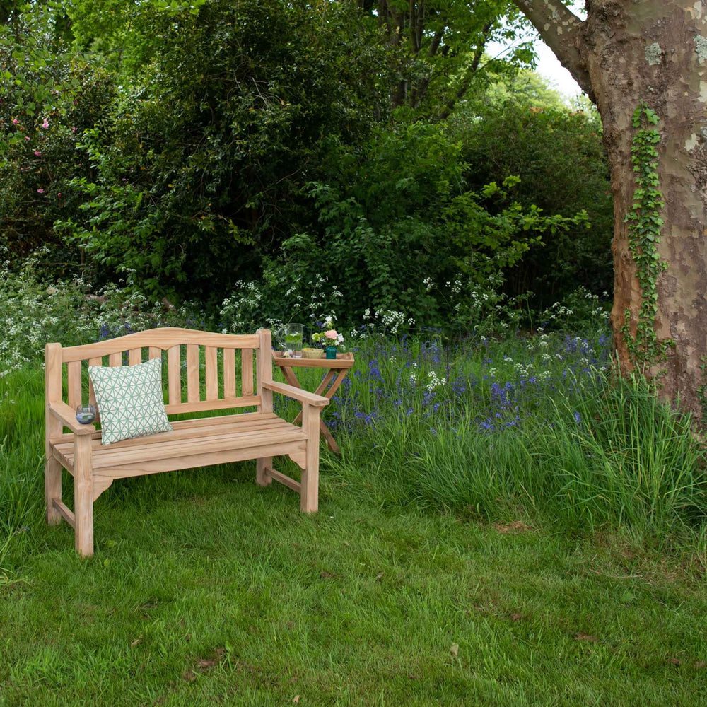 
                  
                    Teak bench with curved backrest and flat armrests, with one green patterned cushion, in grassy garden setting with trees and bushes. 
                  
                