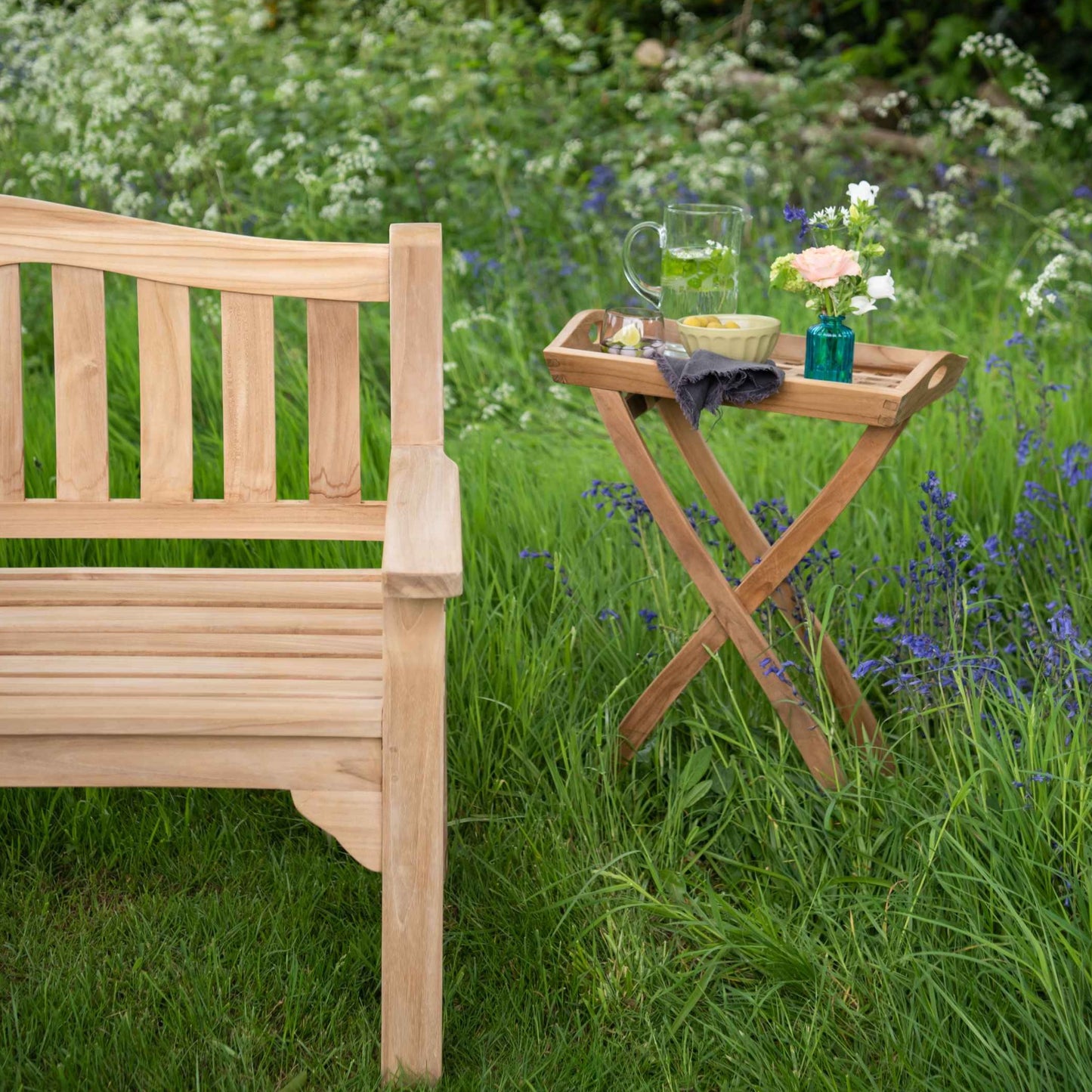 One end of teak bench with curved, slatted backrest and straight flat arms, in lush garden setting with wild flowers.  Next to it is teak butler's tray with stand, holding water jug, bowl and small vase. 