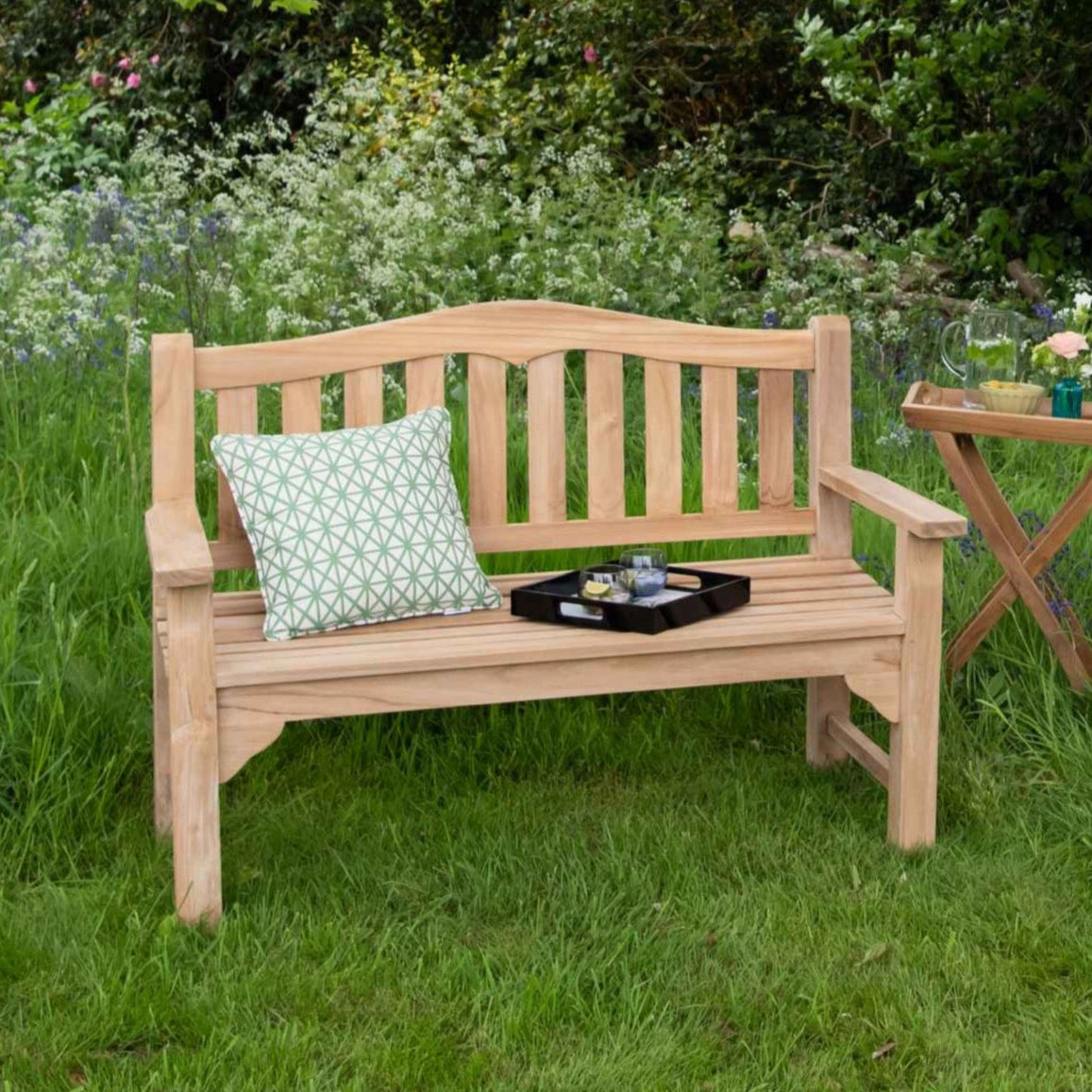 A teak garden bench with a slightly curved slatted seat and backrest, placed on a grassy area. It features flat armrests and is accompanied by a cushion and a small teak folding table with a book and a cup.