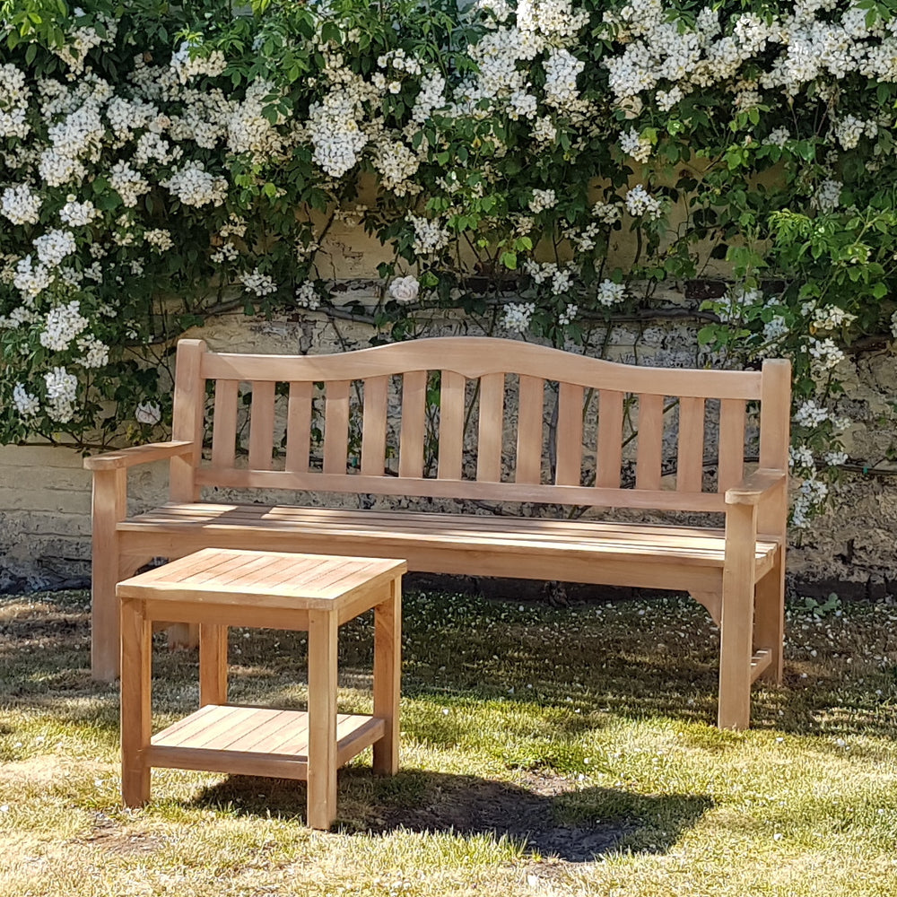 
                  
                    Teak bench with curved backrest and wide slats. In front is a small square teak table. Behind is a stone wall covered in flowers. 
                  
                