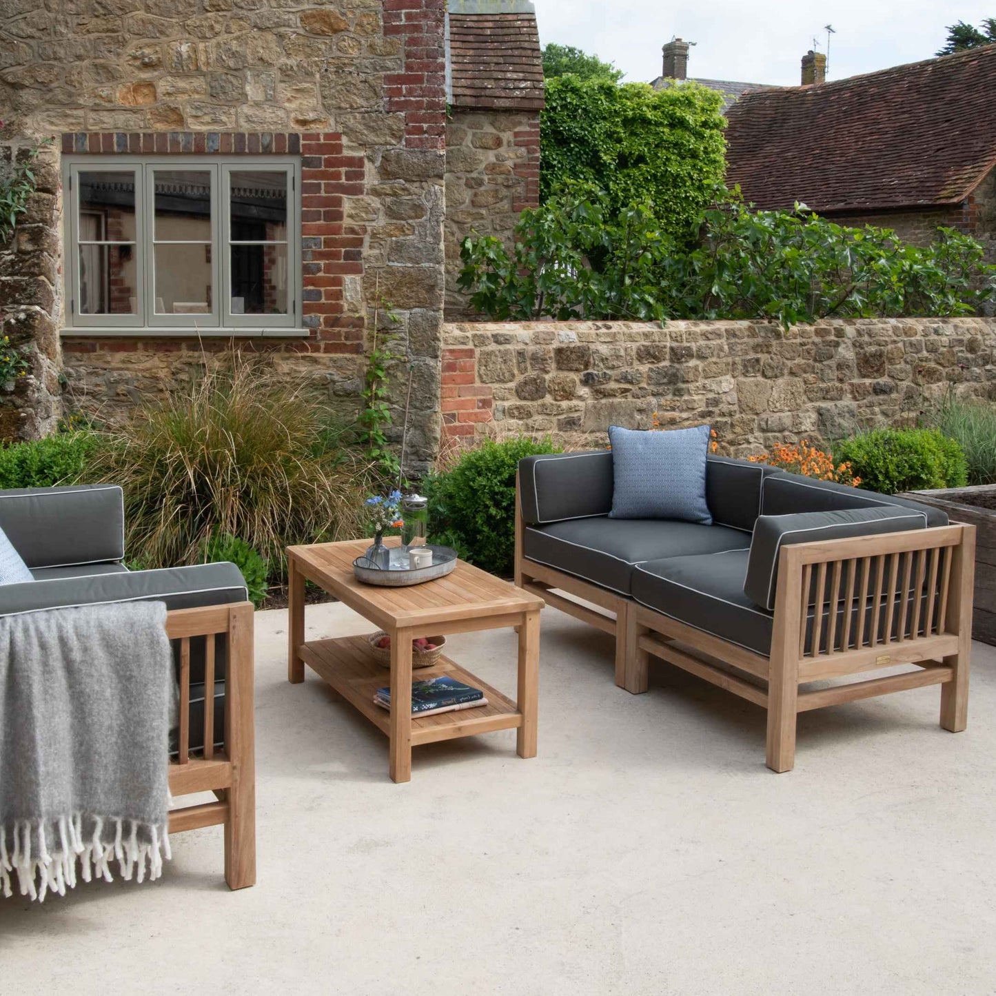 Teak rectangular coffee table on stone patio between two teak benches with armrests and green cushions.On the table is a tray with flowers, and behind is a stone wall and stone and brick house. 